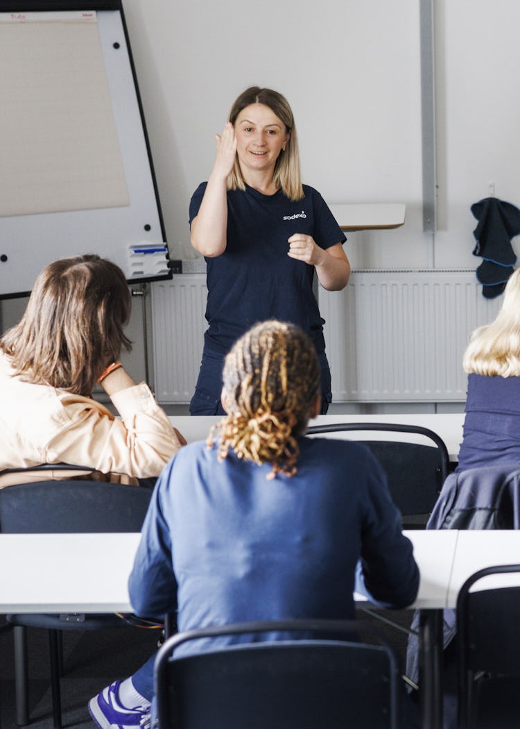 Eldana Hajdari står framför ett klassrum och gestikulerar medan hon talar till en grupp sittande personer. I bakgrunden syns en whiteboard och ett blädderblock.