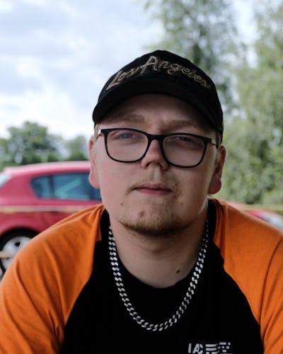 En person med glasögon och en "Los Angeles"-keps bär en orange och svart tröja samt ett kedjehalsband, sittande utomhus med träd och en röd bil i bakgrunden.