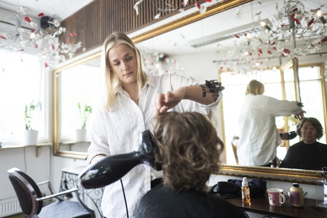 En kvinna i vit skjorta blåser torrt en kunds hår i en salong.