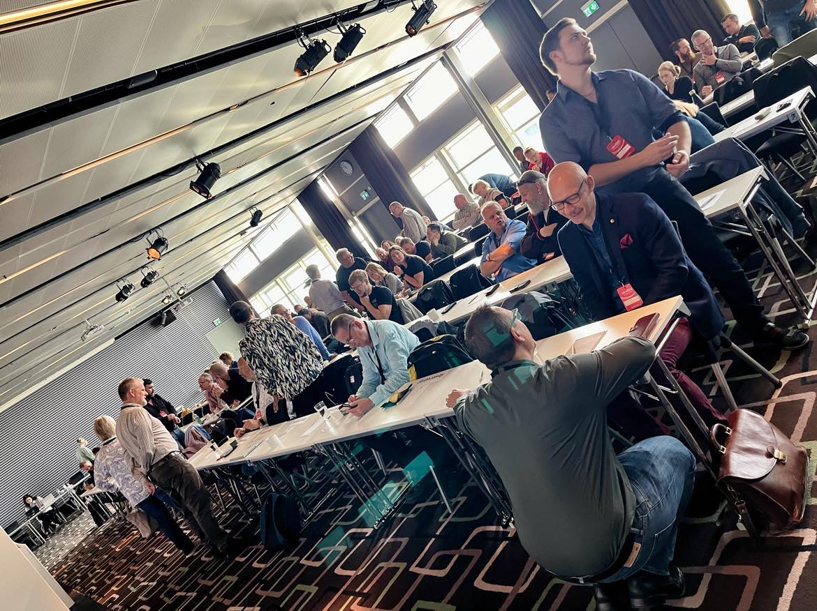 En stor grupp människor sitter vid bord i ett konferensrum och samtal. Vissa personer står upp, och rummet är belyst av naturligt ljus från fönstren.