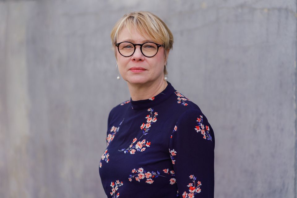 Elisabet Mossberg med kort blont hår och glasögon som bär en marinblå blom-mönstrad topp, står mot en enkel grå bakgrund.