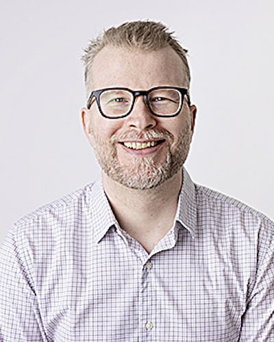 Marcus Nohlberg med glasögon och skägg ler mot kameran. Han har på sig en rutig skjorta. Bakgrunden är vit.