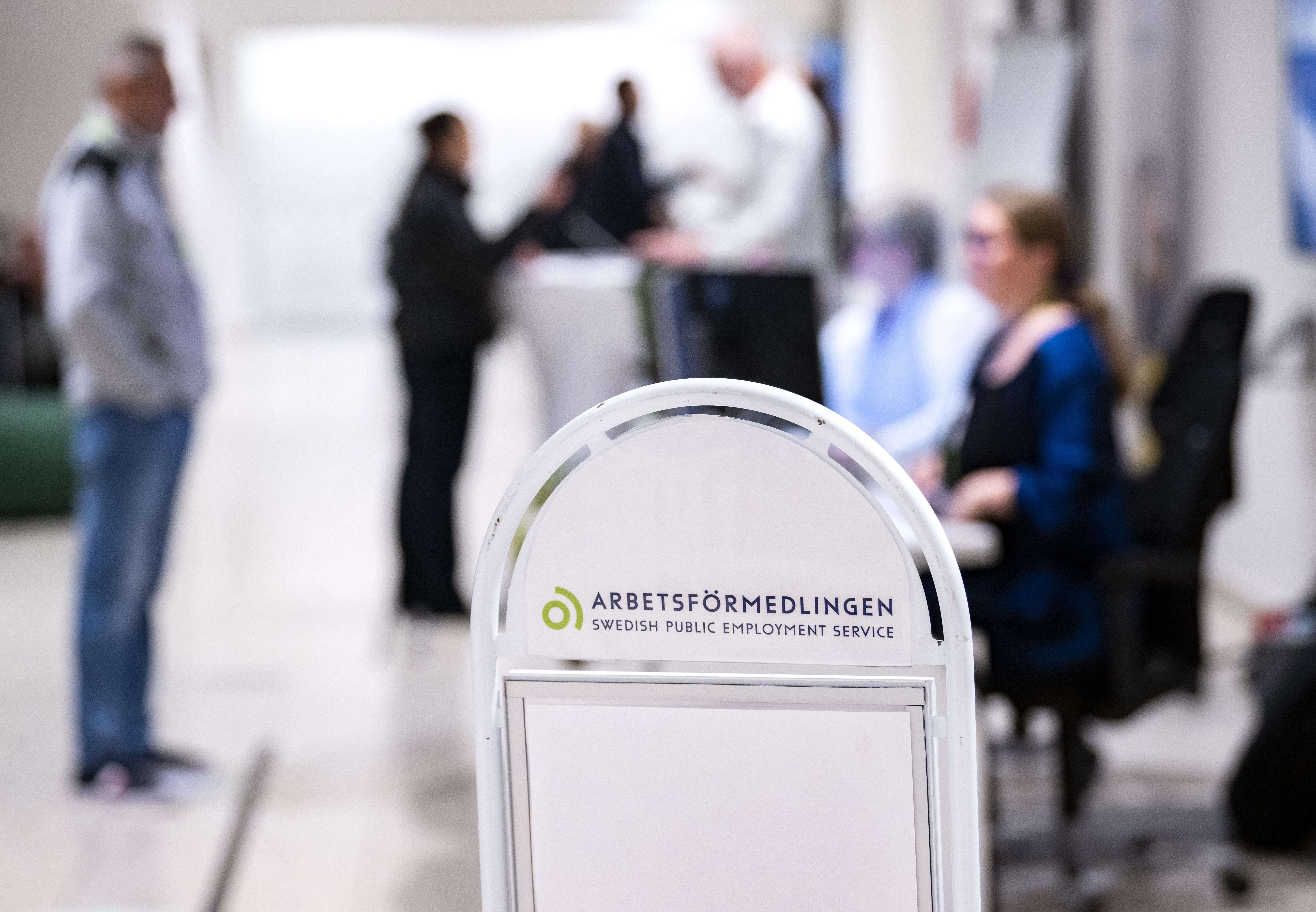 En skylt för Arbetsförmedlingen, den svenska offentliga arbetsförmedlingen, i fokus med människor som interagerar i bakgrunden.