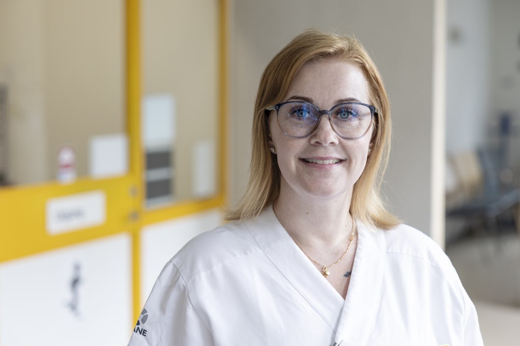 Åza Lundqvist i vit labbrock och glasögon står och ler i en vårdmiljö med gula och vita dörrar i bakgrunden.