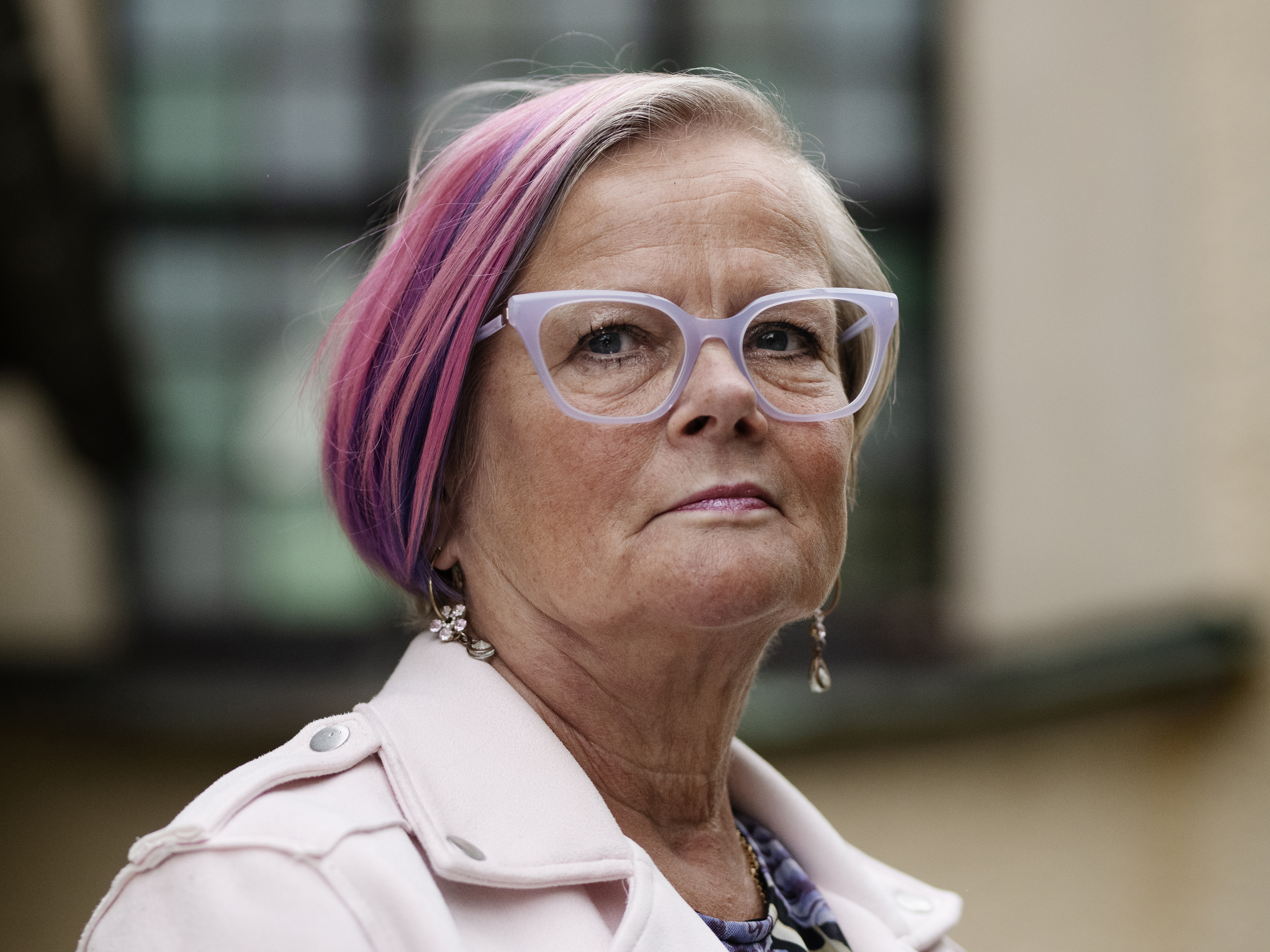 Ewy Gaude med kort lila och rosa hår, vita glasögonbågar och en ljusrosa kappa står utomhus. Hon tittar rakt in i kameran med ett neutralt ansiktsuttryck.