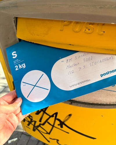 En hand som håller ett blått 2kg paket adresserat till "TM Sweden, Brevlåda 3001, 112 + 3, STOCKHOLM" och placerar det i en gul brevlåda.