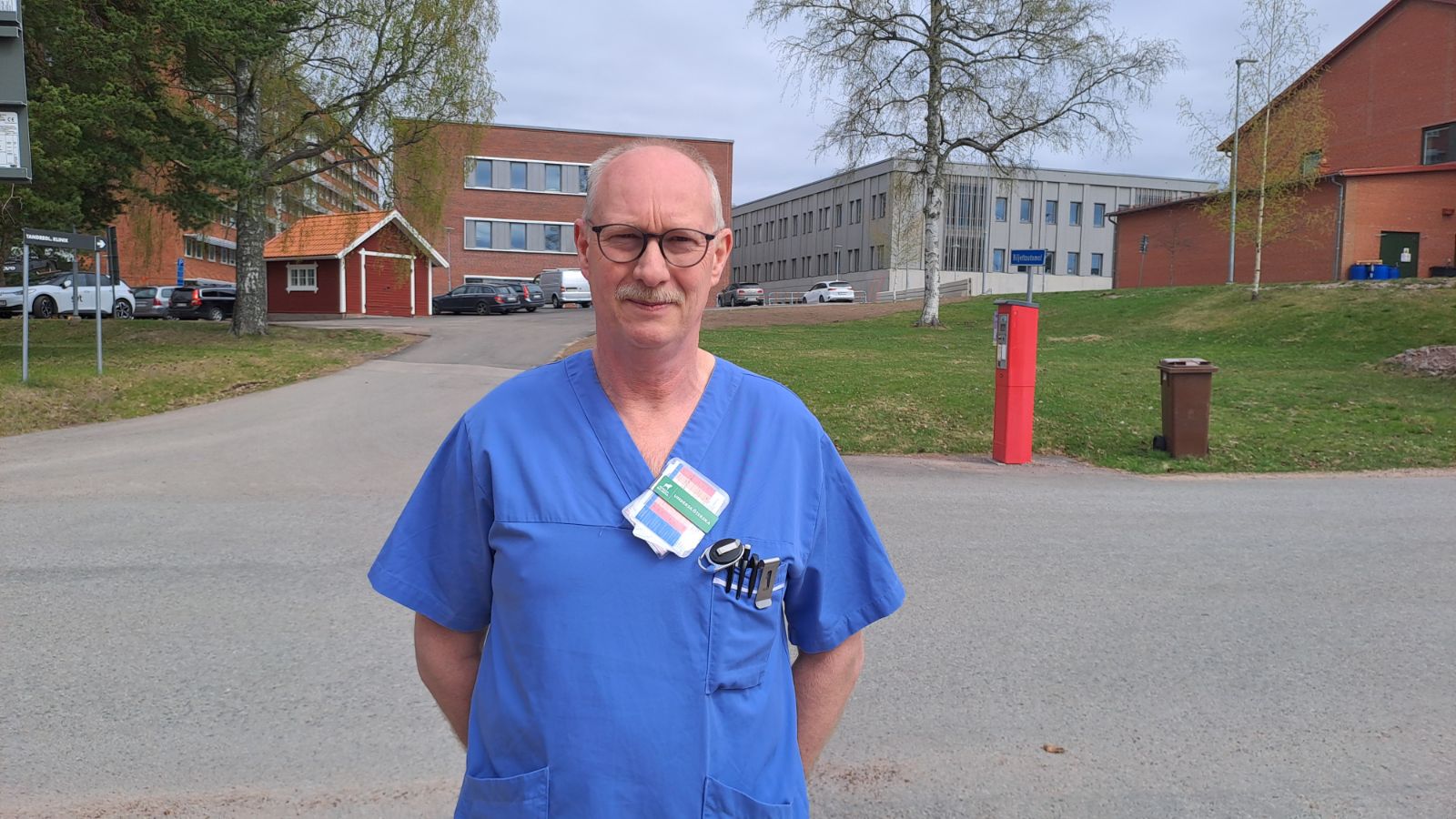 Dick Blaauboer i blå sjukhuskläder står utomhus framför ett sjukhus och andra byggnader, med glasögon, en namnskylt och en penna i bröstfickan. Träd och en väg syns i bakgrunden.