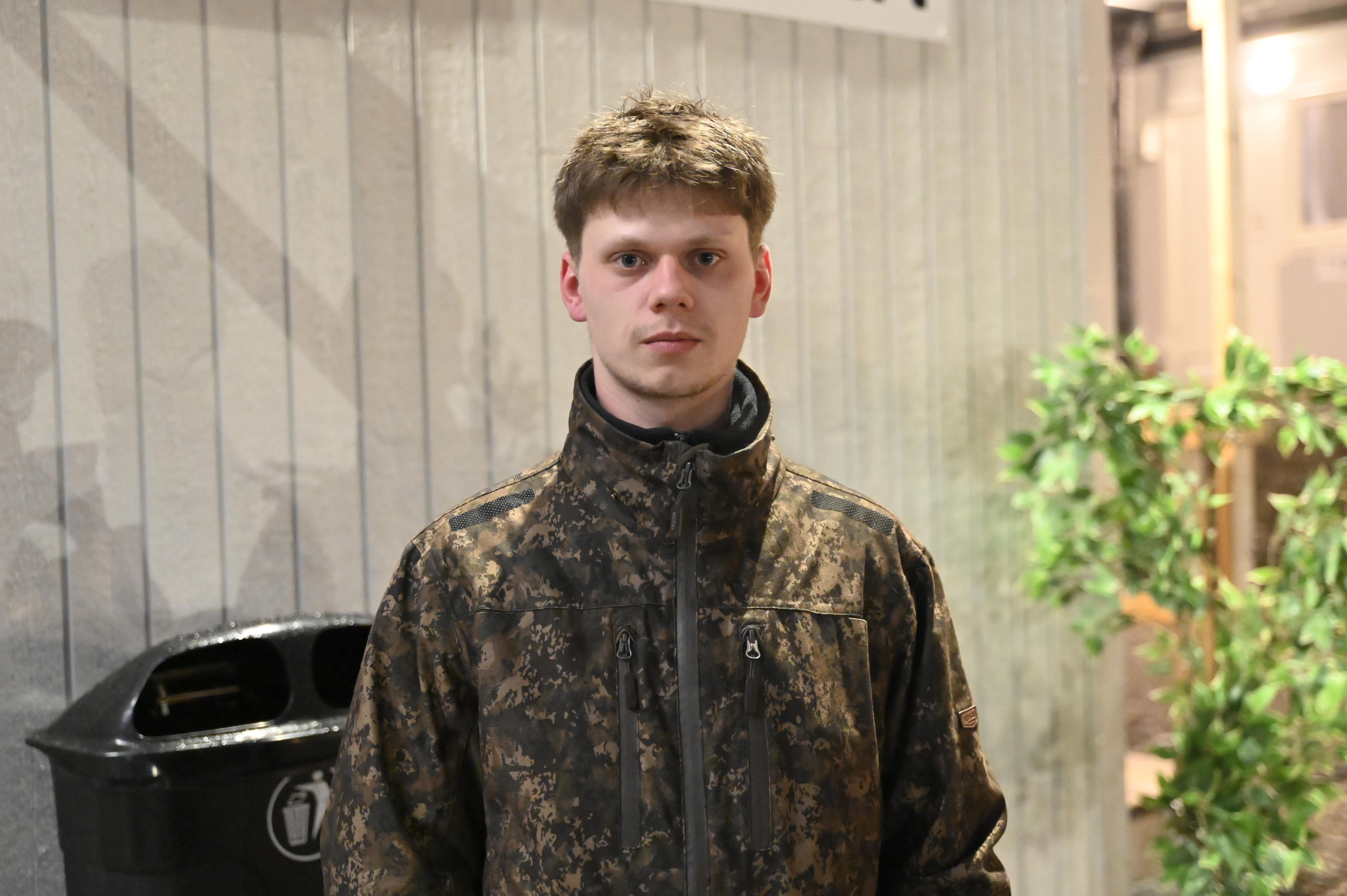 En person i kamouflagejacka står framför en ljus vägg.