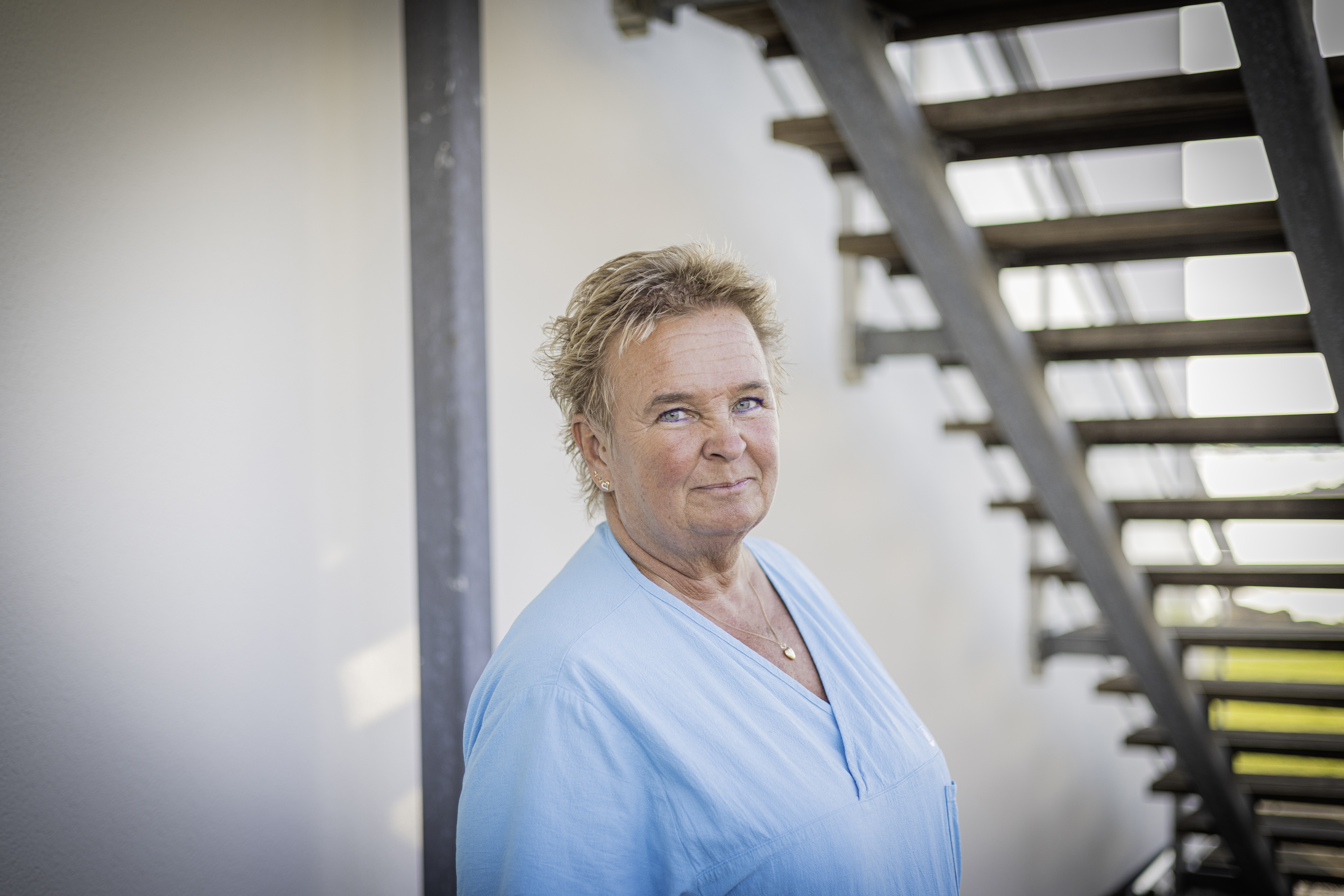 Titti Eriksson, med kort hår och ljusblå skjorta, står nära metalltrappan utomhus och tittar in i kameran.
