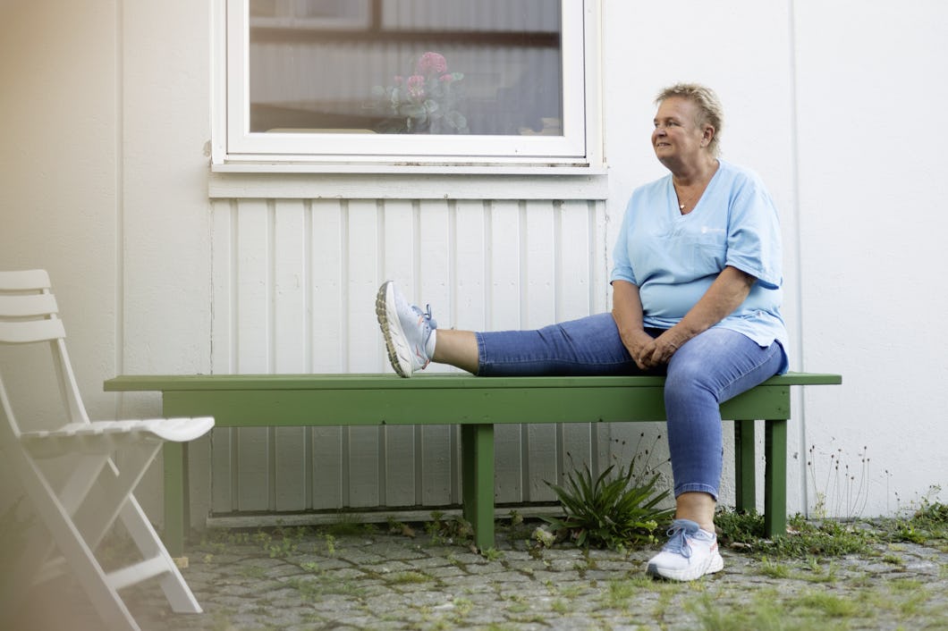 Titti Eriksson med en ljusblå skjorta och jeans sitter på en grön bänk utanför en byggnad, med ena benet utsträckt och det andra böjt. Hon tittar åt sidan utan att visa någon särskild uttryck.