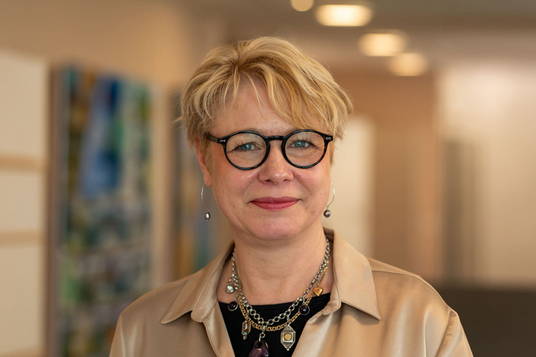 Elisabet Mossberg med kort blont hår och glasögon står inomhus, iklädd en beige blus och ett halsband. Bakgrunden är något suddig.