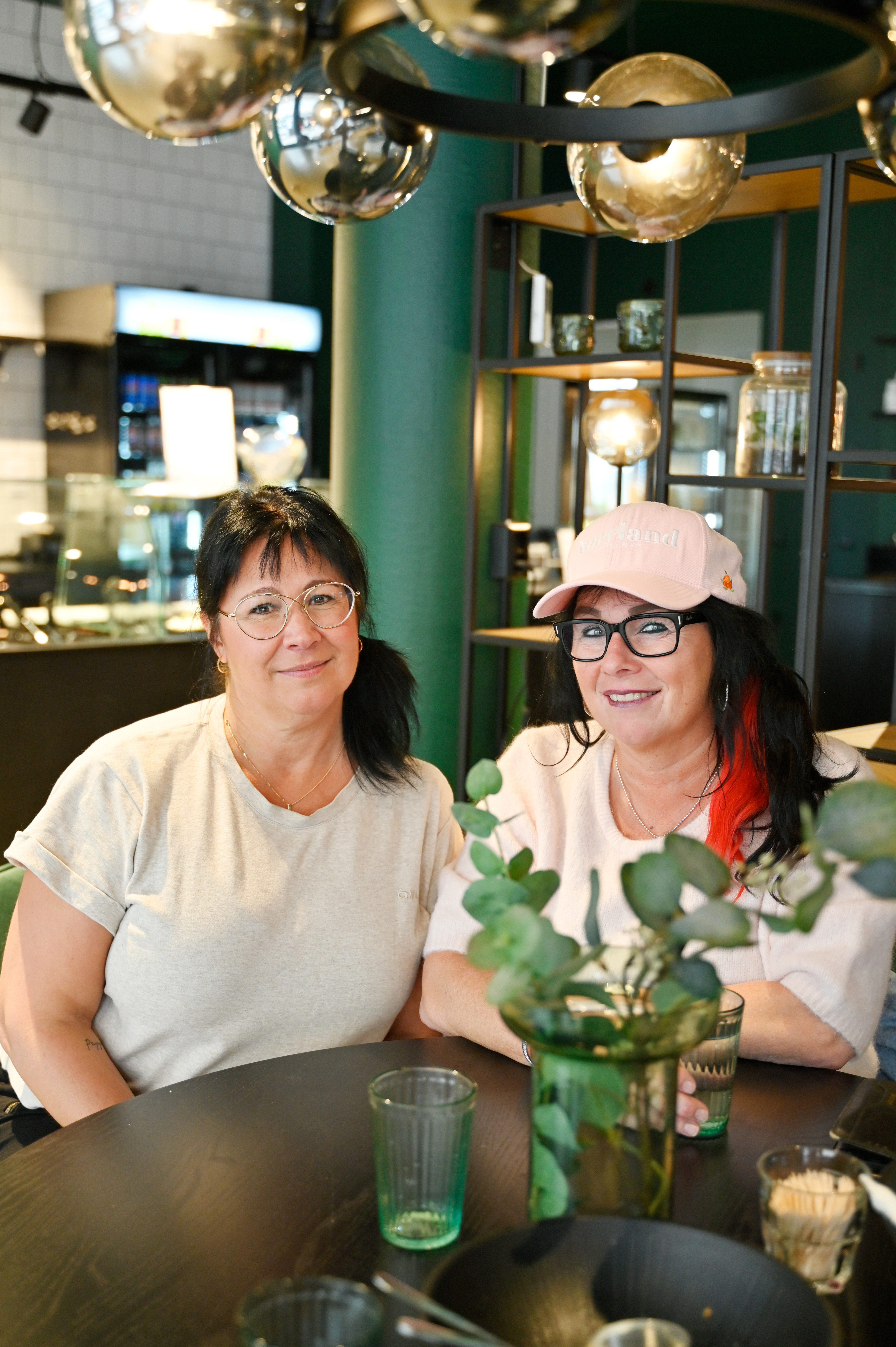 Två kvinnor sitter vid ett runt bord på ett café och tittar in i kameran. De har avslappnade kläder och glasögon. I bakgrunden syns en hylla med varor och en disk.