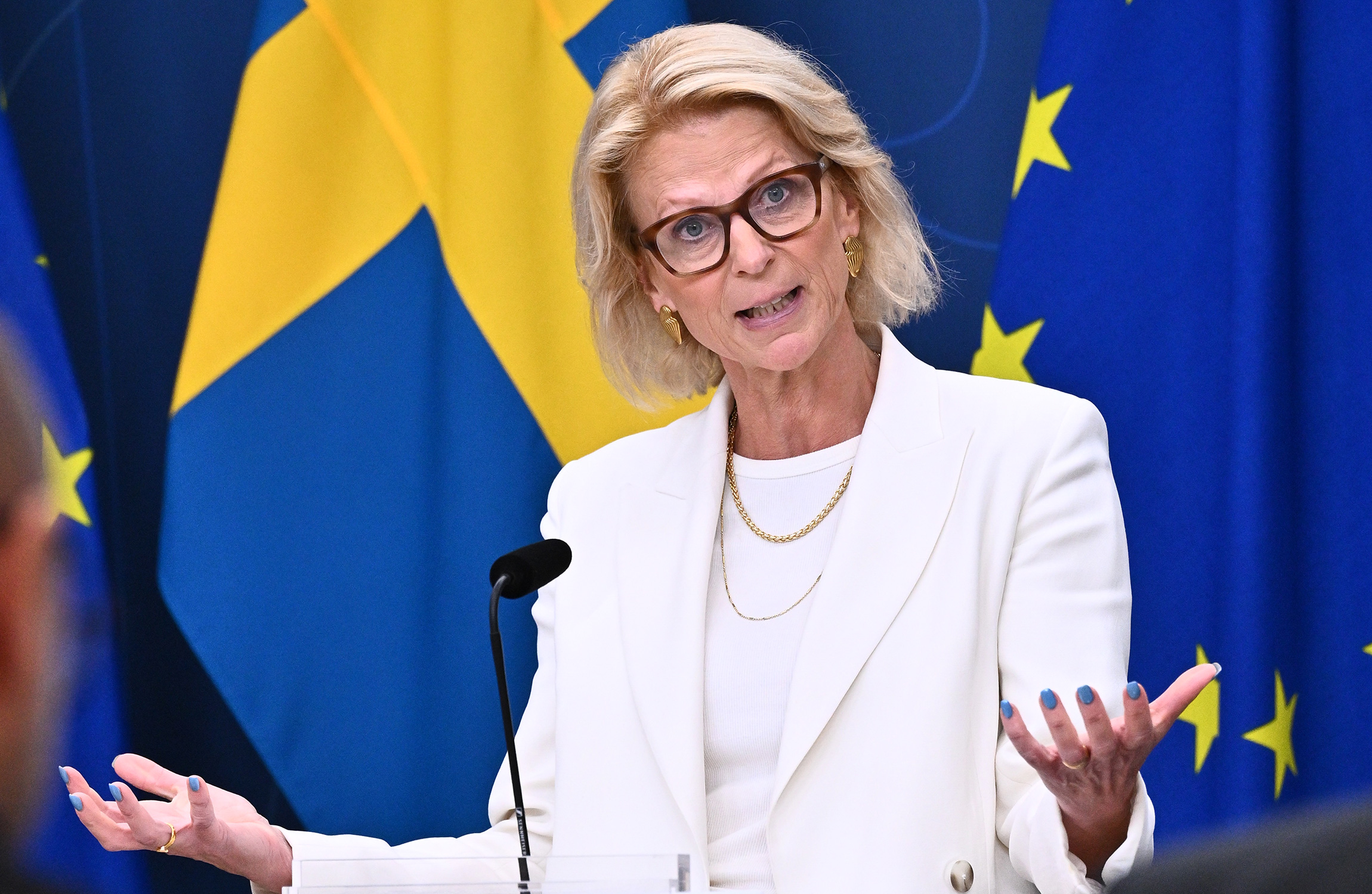 En kvinna i vit kostym och glasögon talar vid ett podium med Sveriges flagga och Europeiska unionens flagga i bakgrunden.