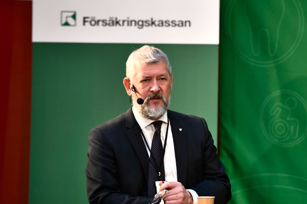 En man i kostym med skägg talar i en mikrofon, stående framför en Försäkringskassan-banner.