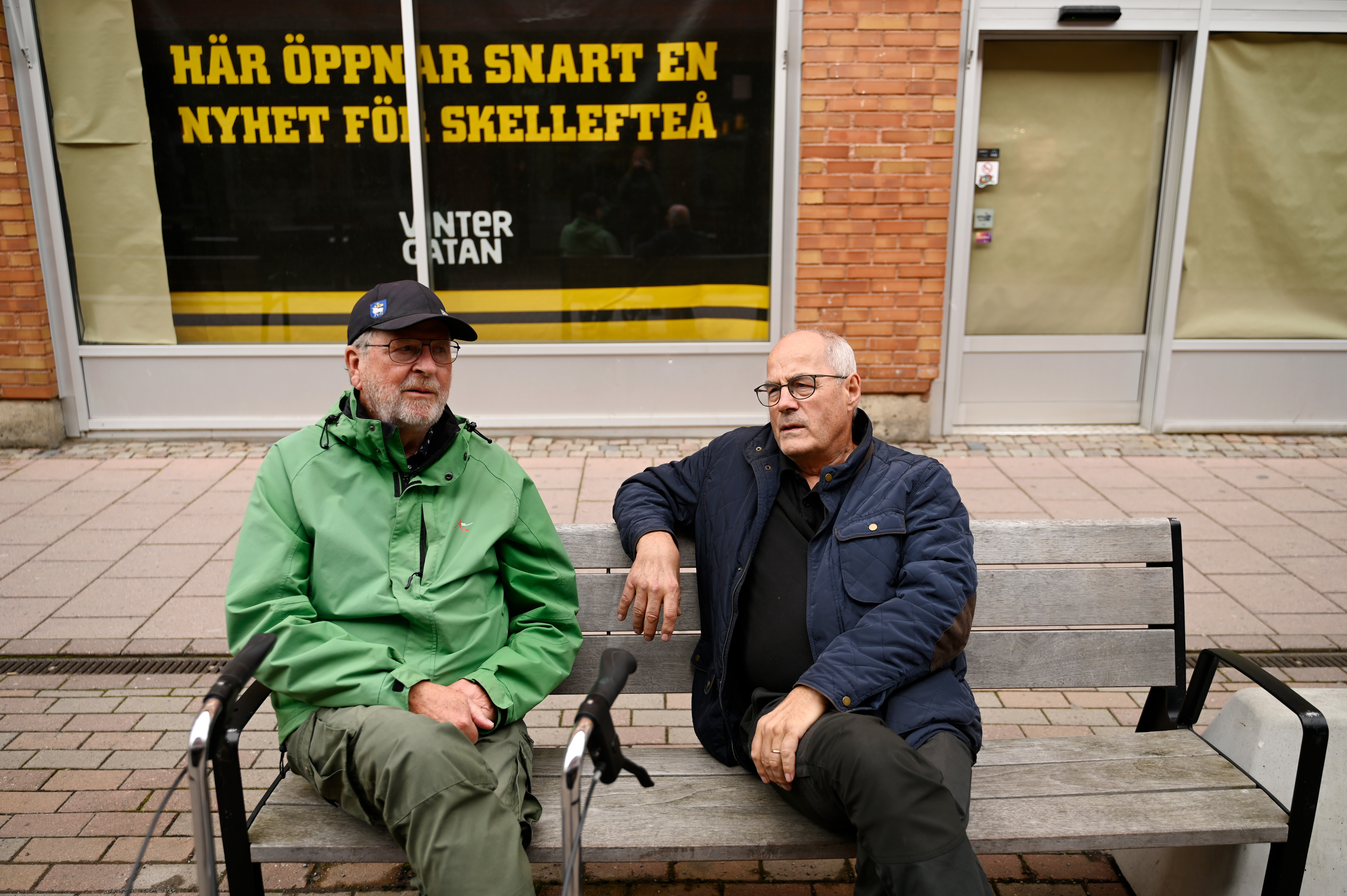 Två äldre män sitter på en bänk och samtalar på en gata. Bakom dem finns en stor skylt på svenska som meddelar att något nytt öppnar snart i Skellefteå.
