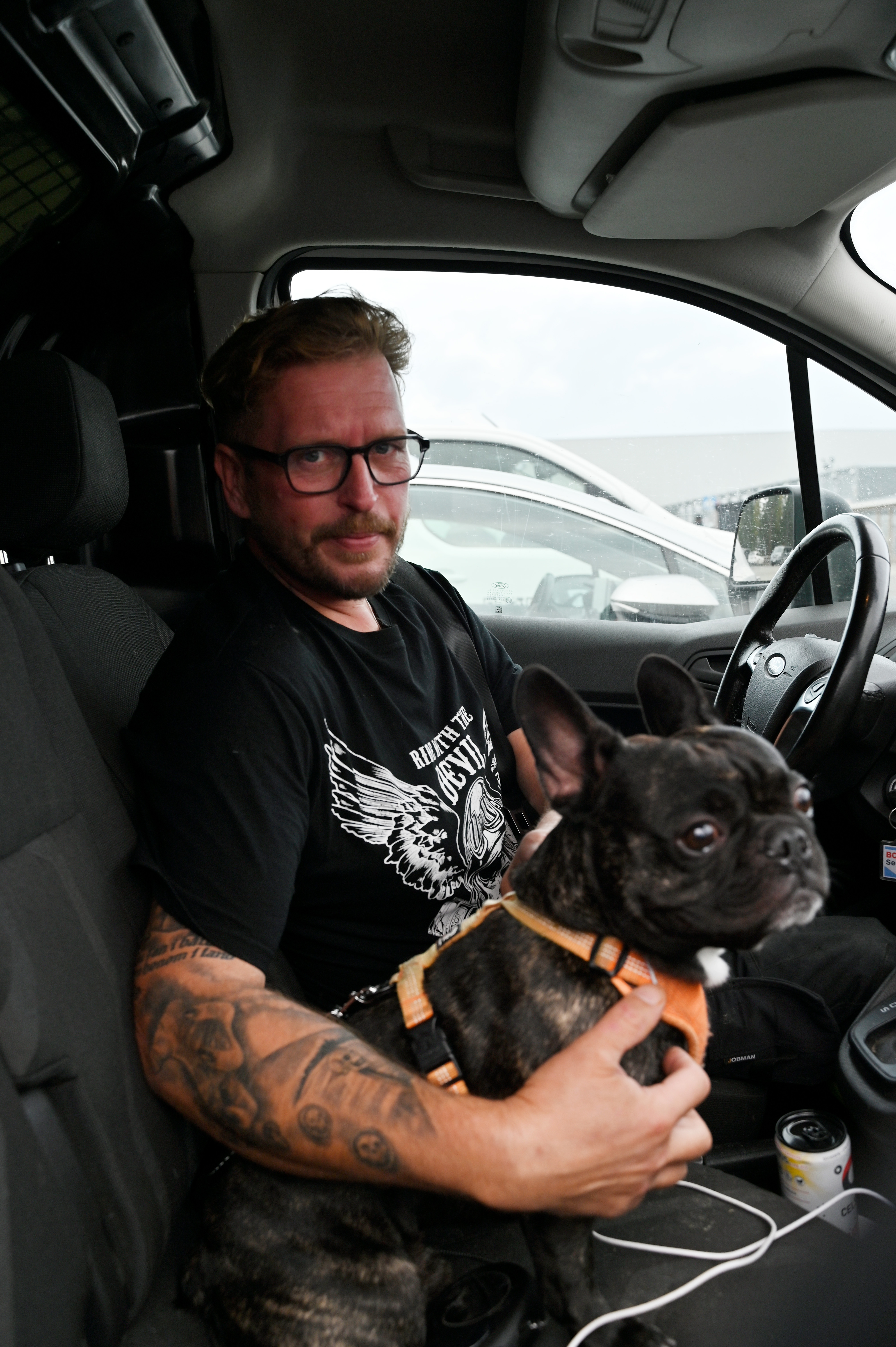 En man med tatueringar och glasögon sitter i en bil med en fransk bulldog i knät.