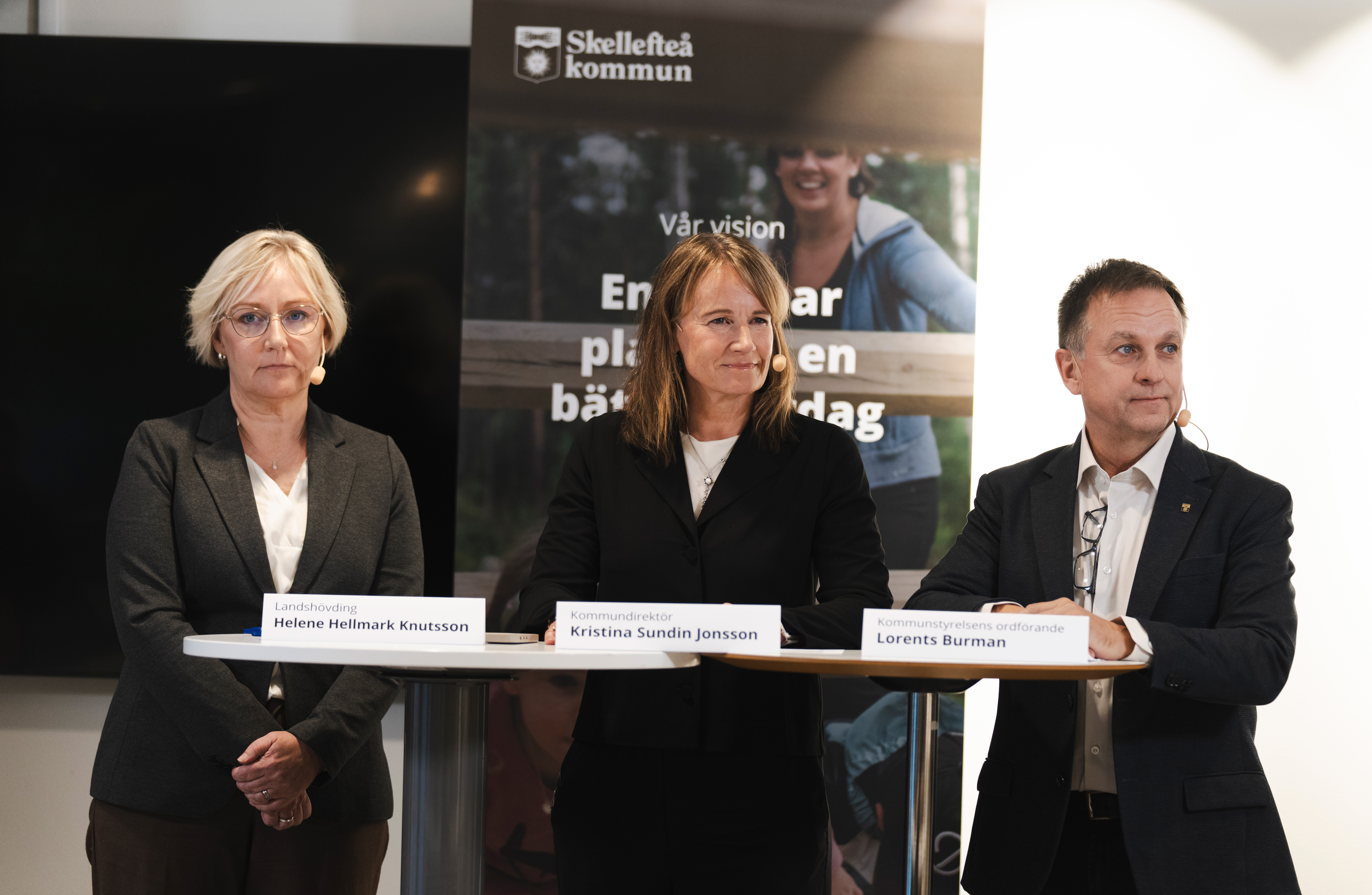 Tre personer står bakom ett bord med namnskyltar vid ett Skellefteå kommun-evenemang. En banner i bakgrunden visar organisationens vision.
