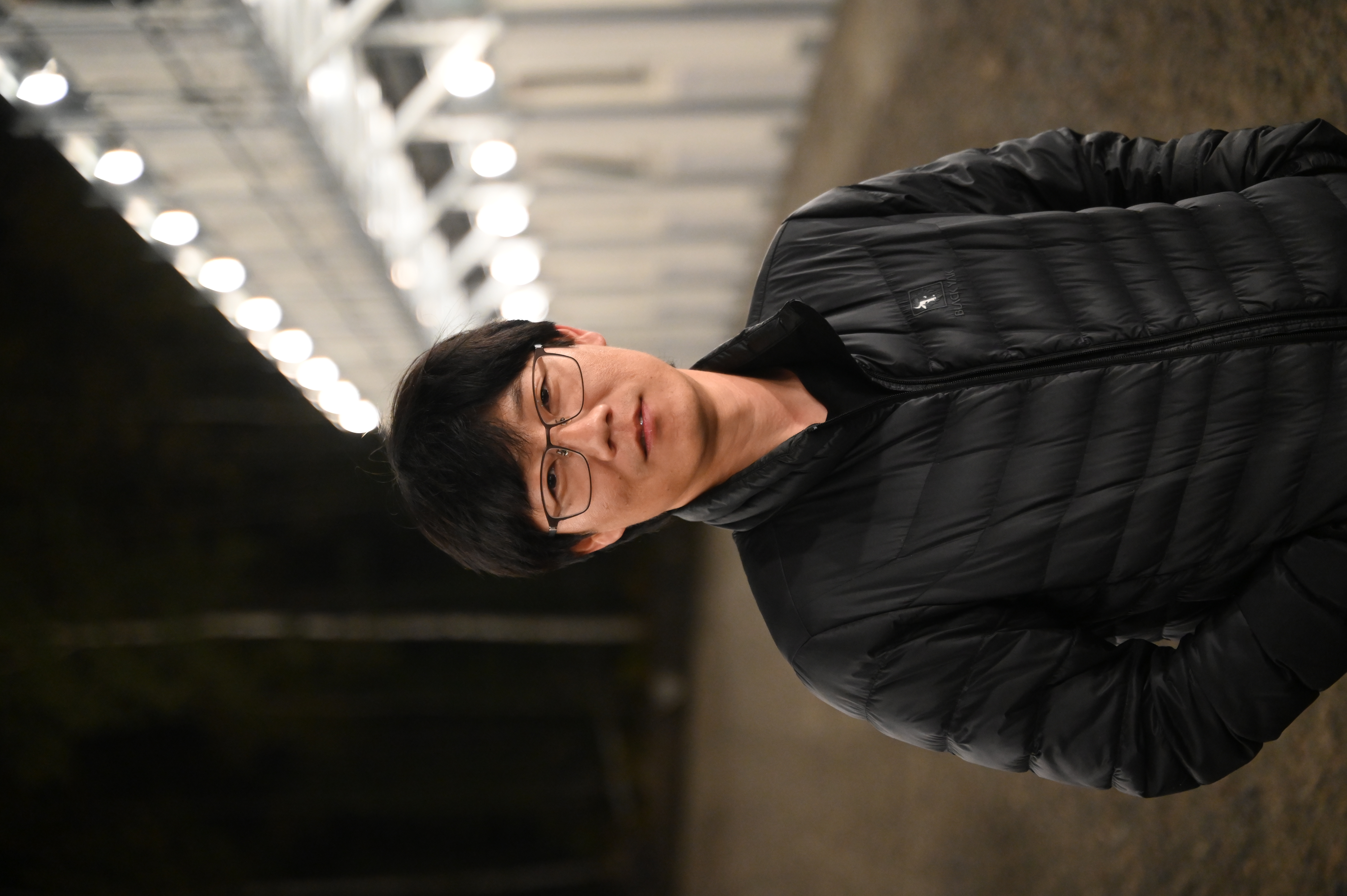 En person med kort svart hår och glasögon står utomhus , iförd en svart dunjacka. I bakgrunden syns en väl upplyst byggnad omgiven av ljus.