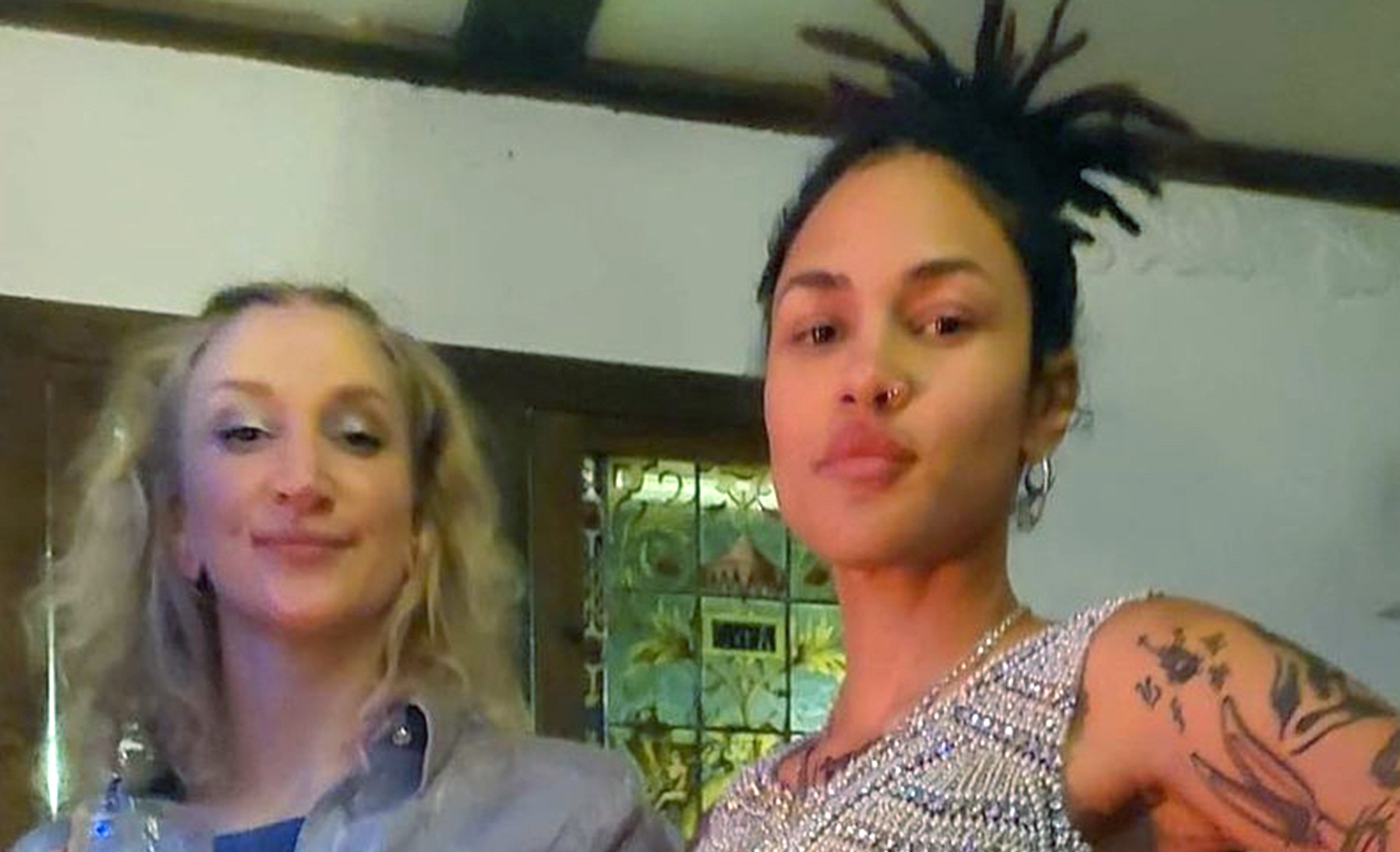 Två personer står inomhus, en med bakåtbundet blont hår och den andra med mörkt hår i dreadlocks. Båda har på sig vardagskläder och tittar in i kameran.