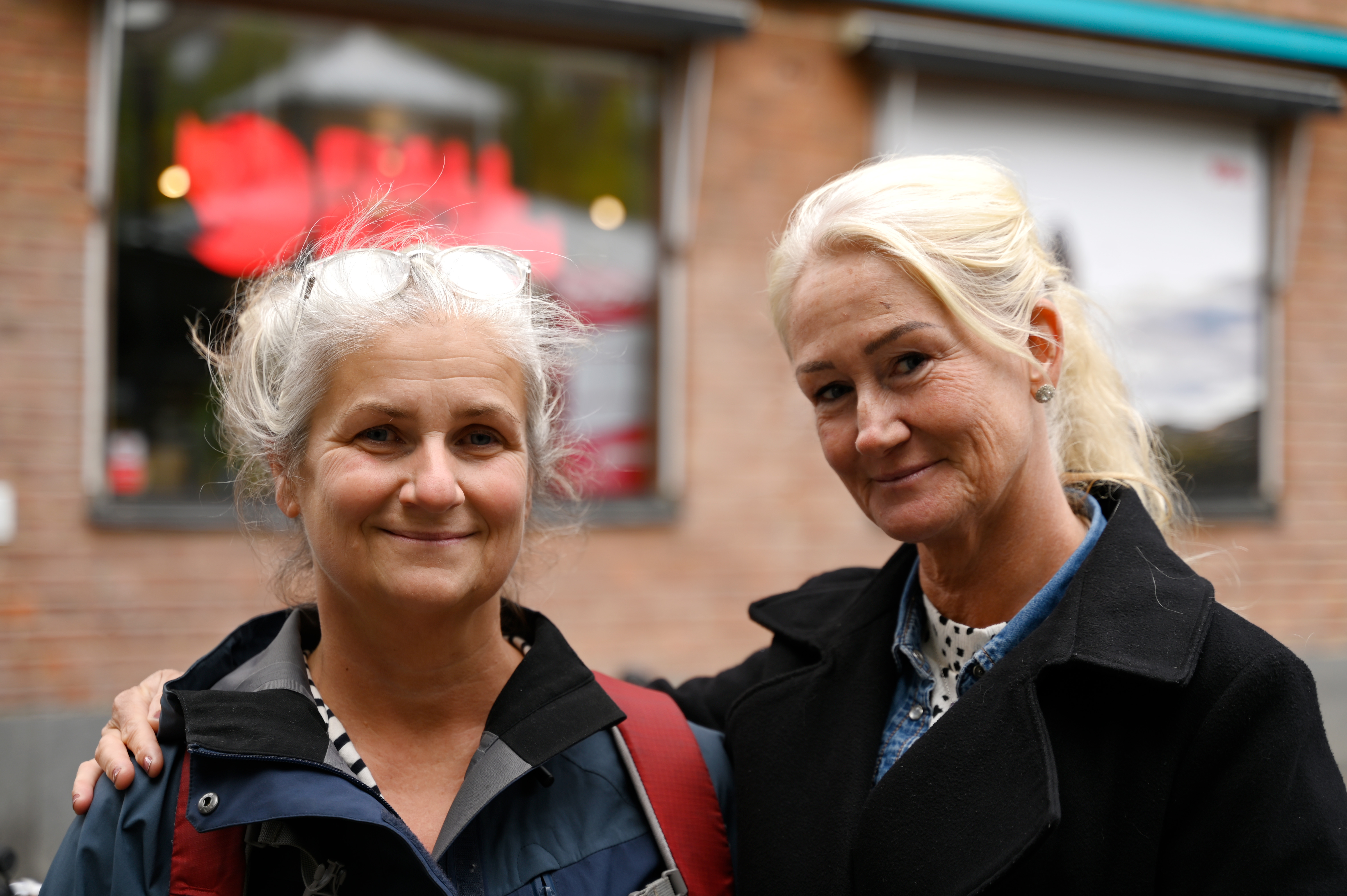 Två kvinnor med grått hår står tillsammans utomhus. Den ena bär glasögon och en jacka, medan den andra har en svart kappa. I bakgrunden syns en tegelvägg och ett skyltfönster.