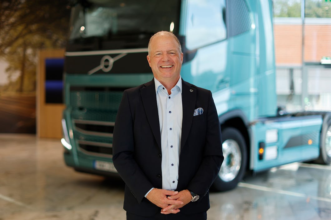 En man i kostym står framför en grön Volvo-lastbil i ett väl upplyst inomhusutrymme.