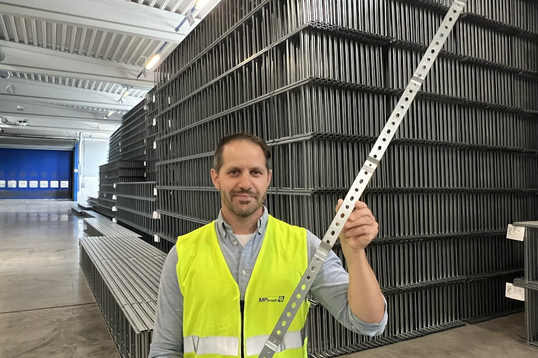 En man i en säkerhetsväst håller i en perforerad metallremsa i ett lager fyllt med staplade metallplåtar.