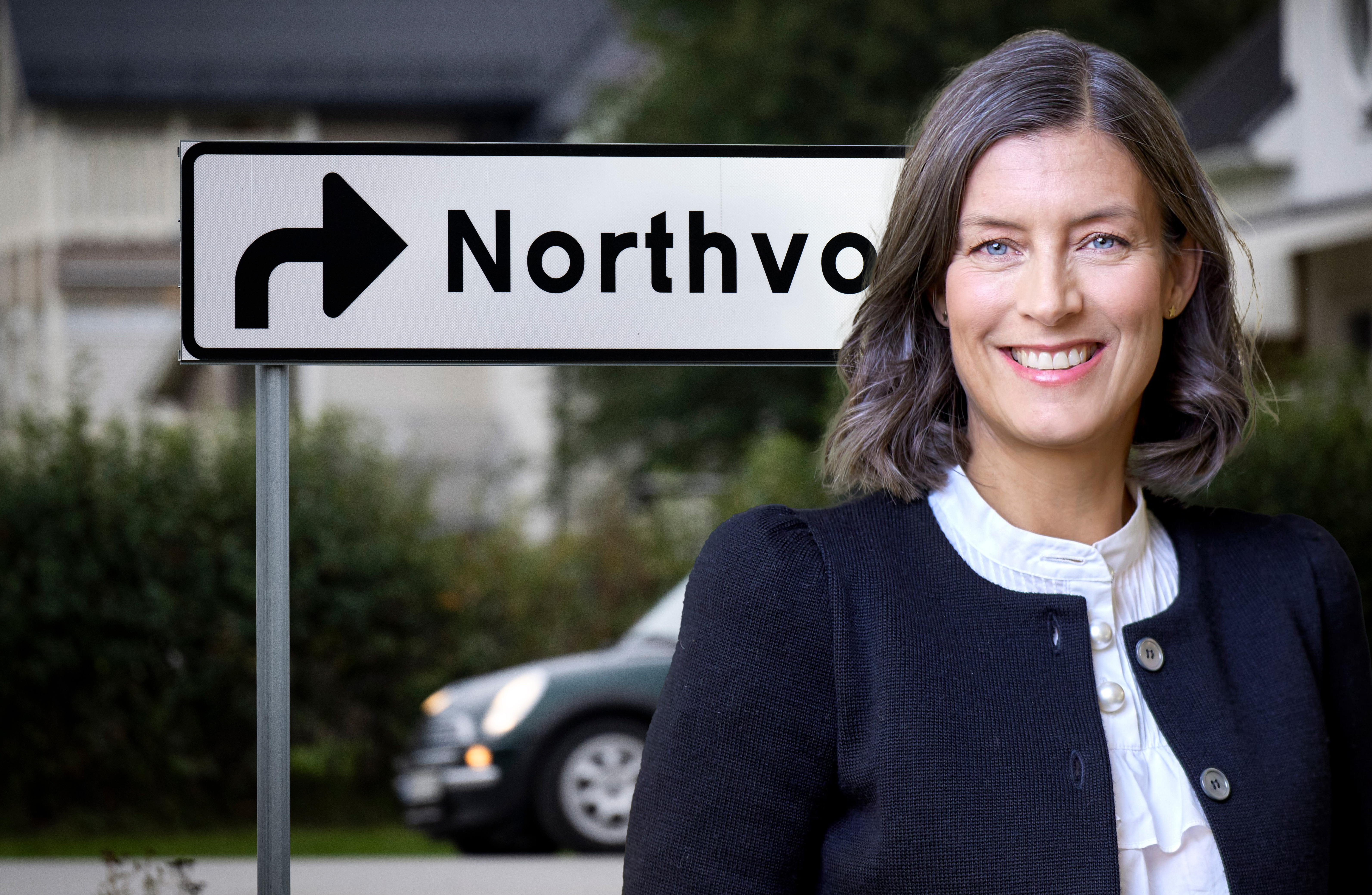 En kvinna med grått hår står framför en vägskylt som har texten "Northvolt" och en pil åt höger. En bil syns i bakgrunden.