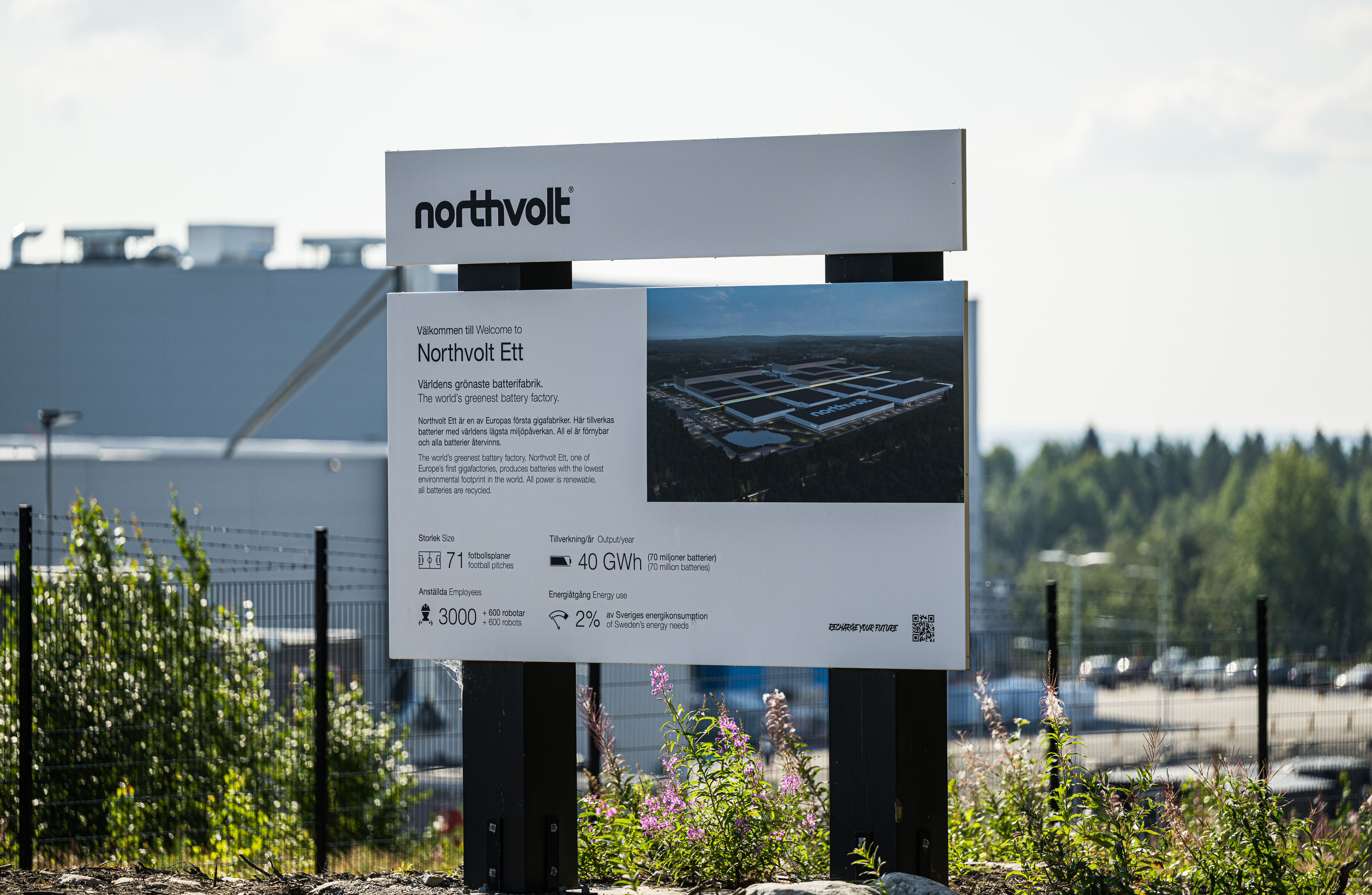En Northvolt Ett-skylt framför en industribyggnad och grönska. Skylten informerar om tillverkningsanläggningen för batterier.