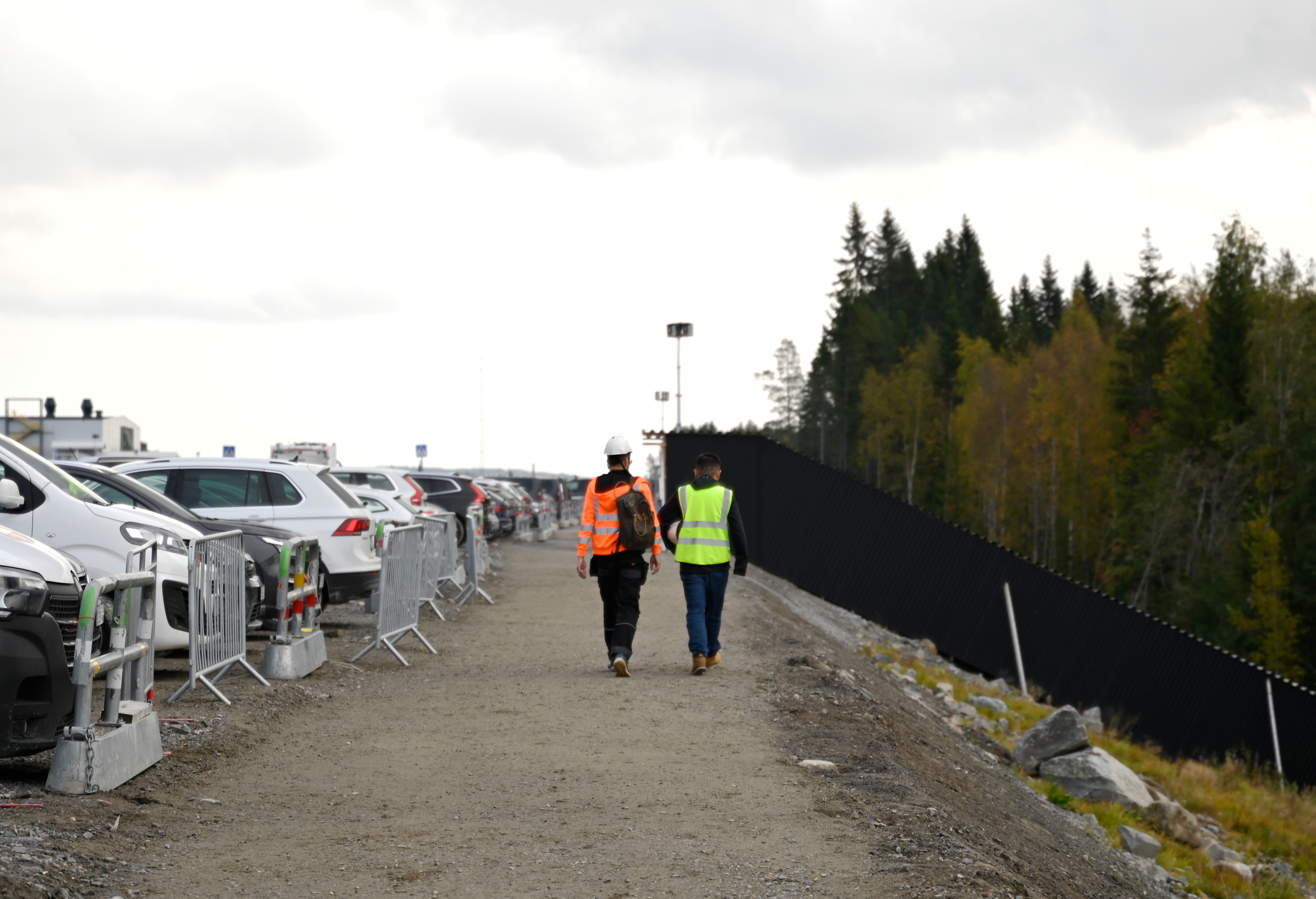 Två byggarbetare med högsynlighetsvästar går längs en grusstig intill parkerade bilar och ett skogsområde.