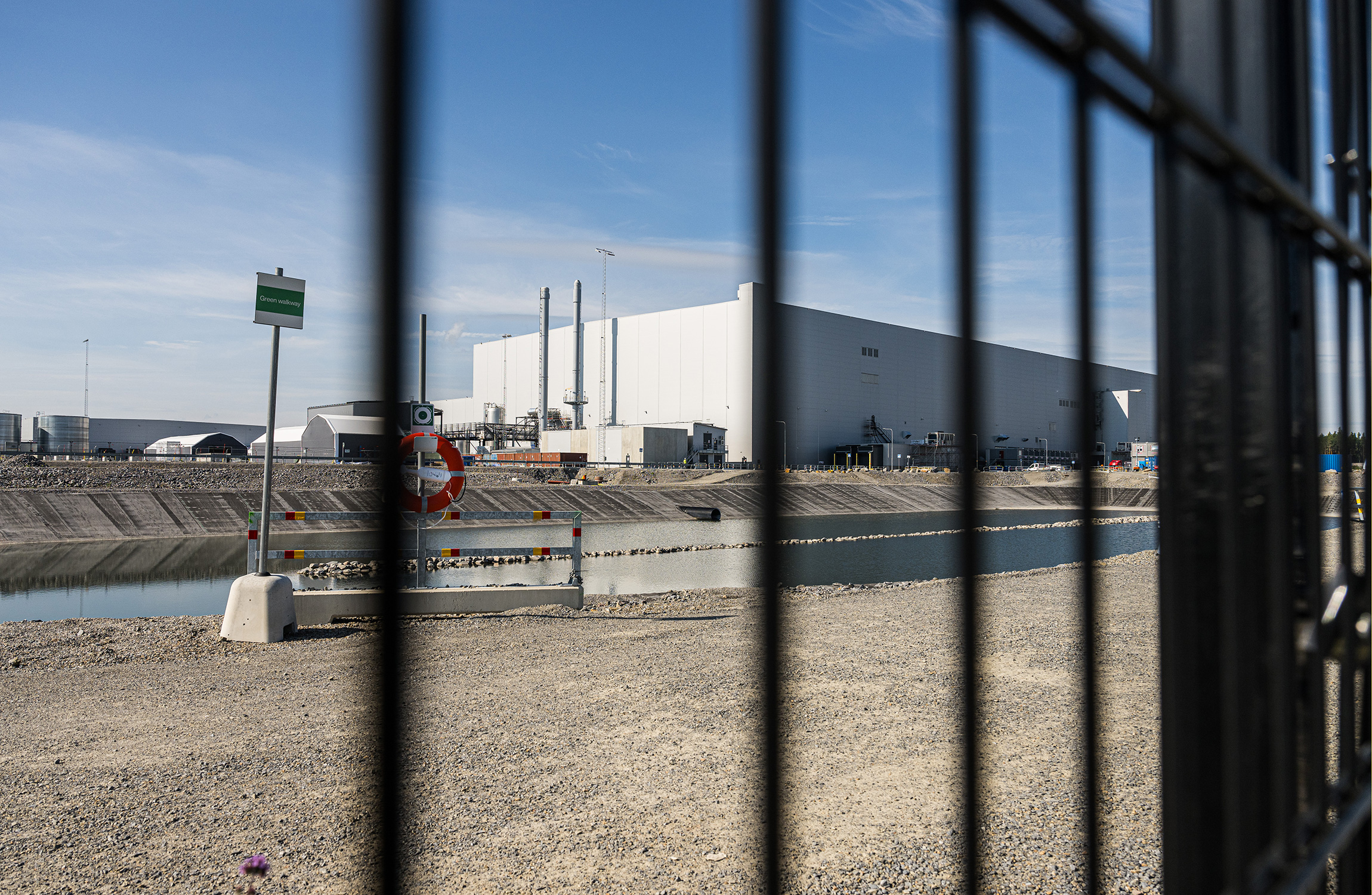 En stor industribyggnad syns tvärs över en vattenmassa, sedd genom ett svart metallstaket på en klar dag.