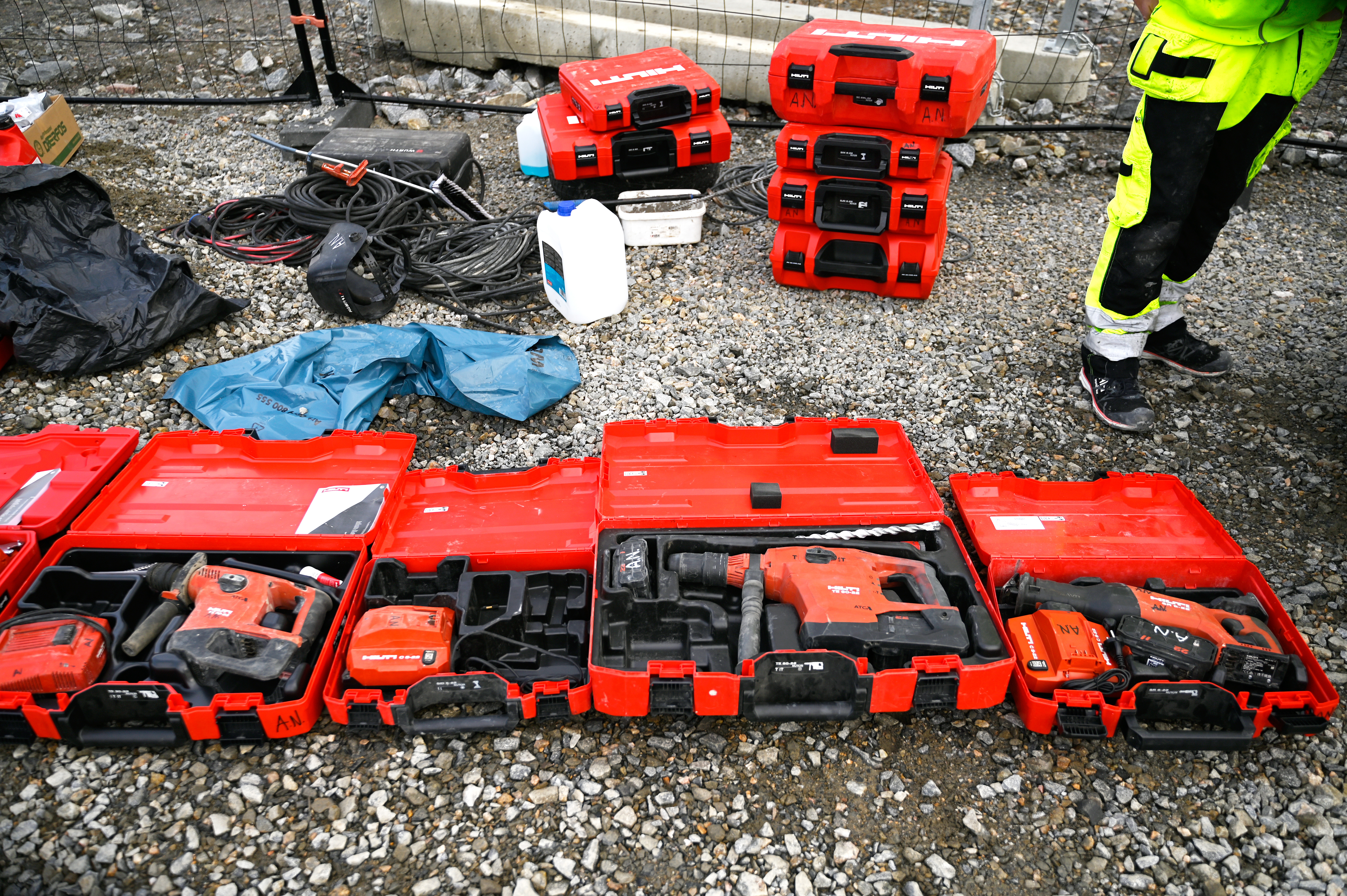 Flera röda verktygslådor som innehåller olika elverktyg är utlagda på grus. I bakgrunden syns fler verktygslådor och byggmaterial. En person i kläder med hög synlighet är delvis synlig.