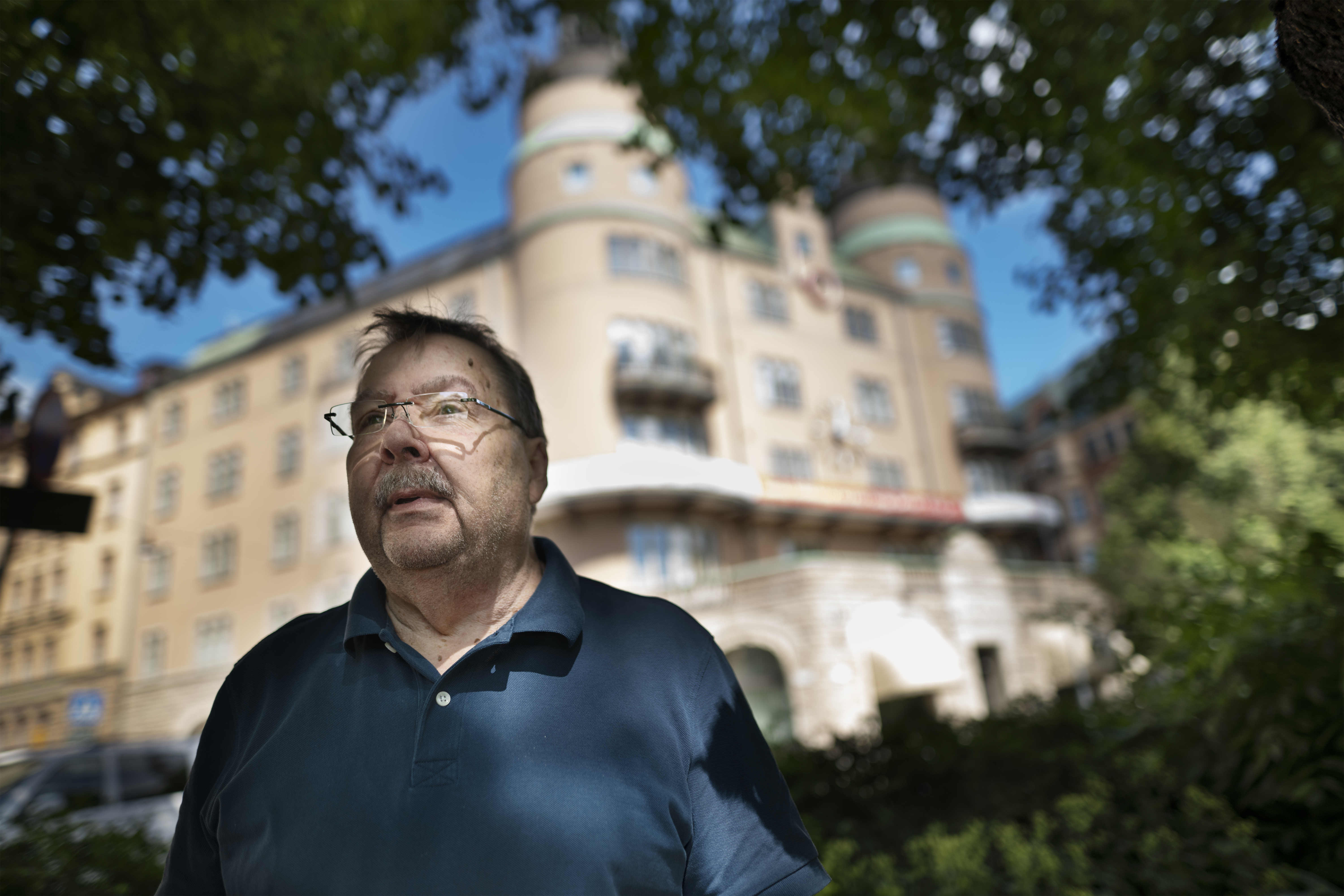 En äldre man med glasögon står utomhus, omgiven av träd. I bakgrunden syns en historisk byggnad med runda torn.
