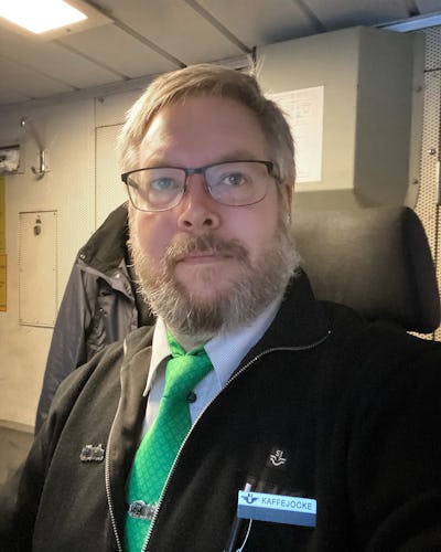 Joakim Thörvall bär en svart jacka, grön slips och en namnbricka som det står "KAFFEJOCKE" på.