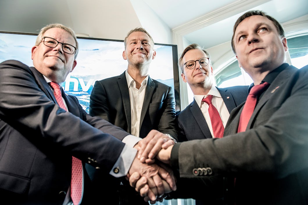 Fyra män i kostymer står nära varandra och skakar hand. De poserar för kameran, ser ut att vara enade och professionella. En skärm är synlig i bakgrunden.