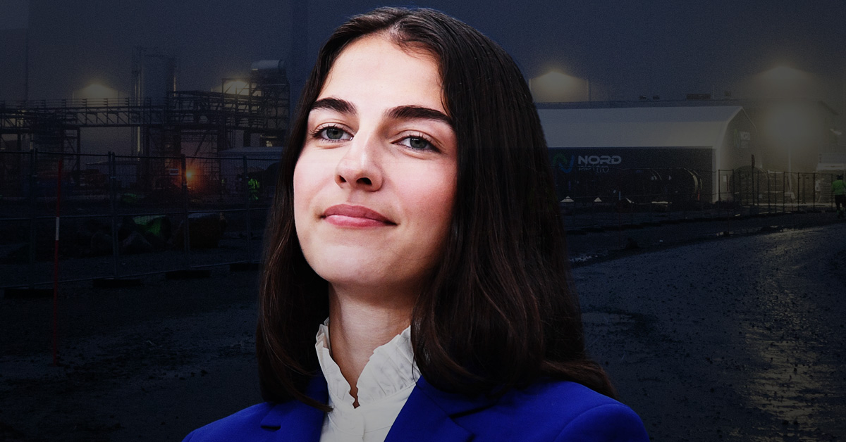 En kvinna med långt brunt hår och blå kavaj står framför en industriplats med dämpad belysning.