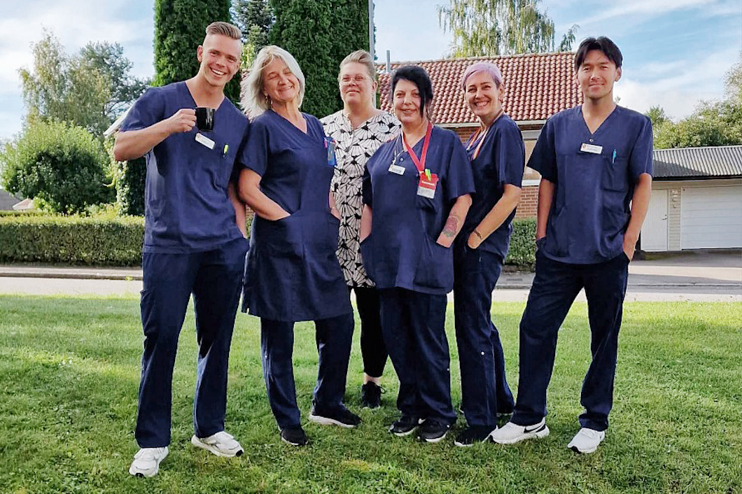 En grupp med fem vårdpersonal i blå arbetskläder och en person i en vit och svart mönstrad klänning står utomhus på ett gräsområde, vända mot kameran.