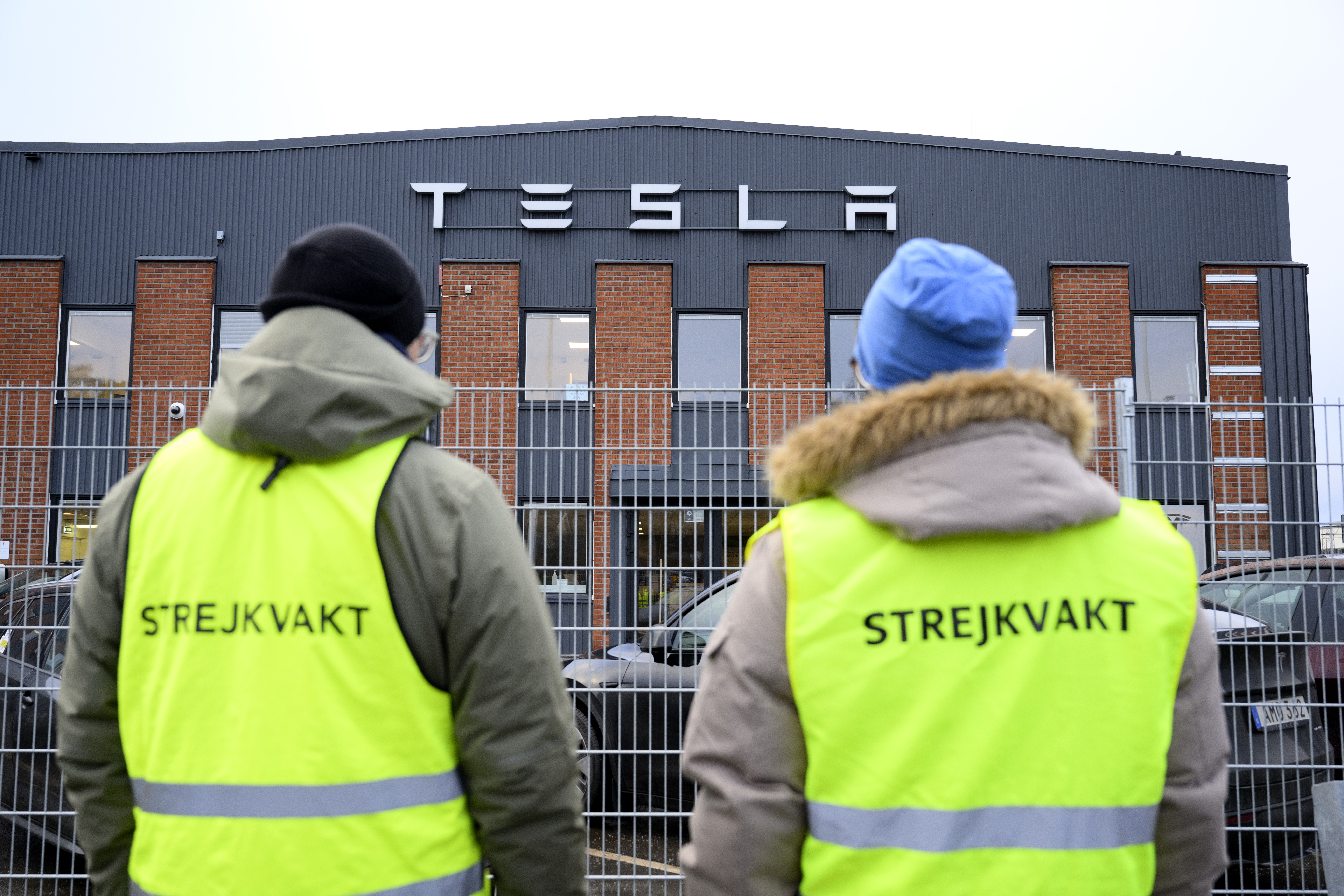 Två personer i gula västar med texten ”Strejkvakt” står utanför en Tesla-byggnad.