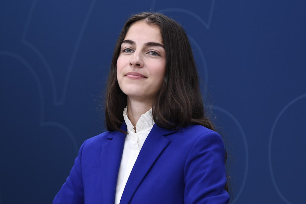 Klimat- och miljöminister Romina Pourmokhtari med långt mörkt hår, klädd i en blå blazer och vit skjorta, står mot en blå bakgrund.