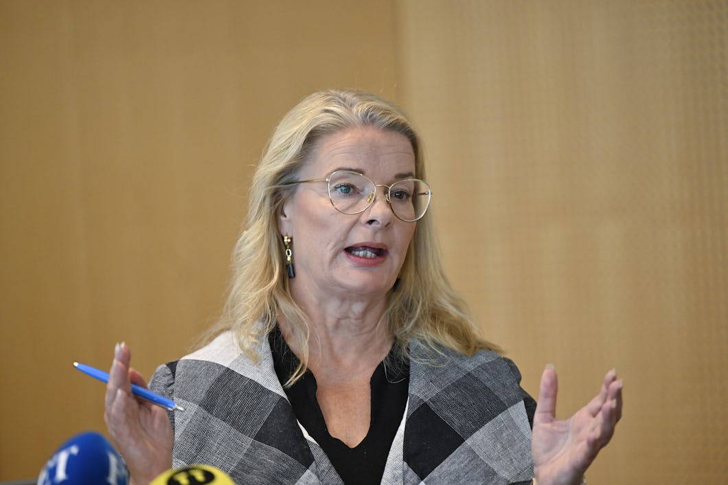 Skolminister Lotta Edholm (L) med glasögon håller en penna och gestikulerar medan hon talar vid en presskonferens. Flera mikrofoner syns i förgrunden.