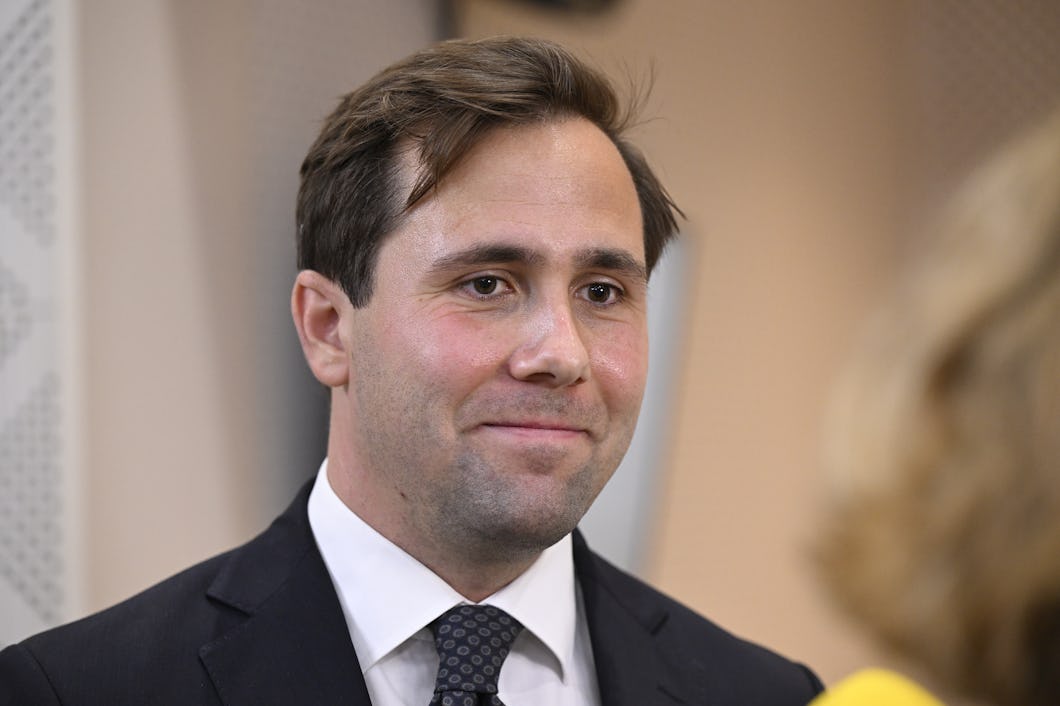 Benjamin Dousa med kort brunt hår, klädd i kostym och slips, tittar något bort från kameran. En person med blont hår och en gul skjorta är delvis synlig i förgrunden.