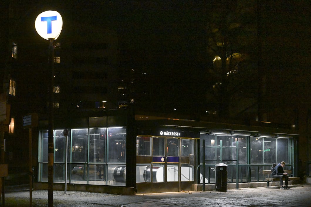 Nattvy av en upplyst tunnelbaneingång med en ”T”-skylt och en person som sitter på en bänk utanför.