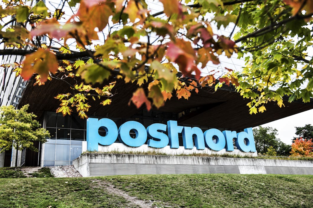 En byggnad med en stor, blå Postnord-logotyp på framsidan, delvis skymd av höstlöv från närliggande träd.