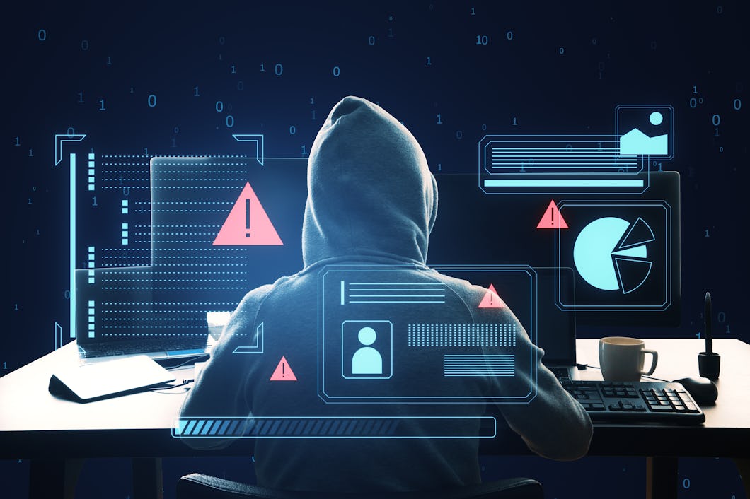En person i en huvtröja sitter vid ett skrivbord med flera datorskärmar som visar grafik och varningssymboler, vilket representerar cybersäkerhetsshot.