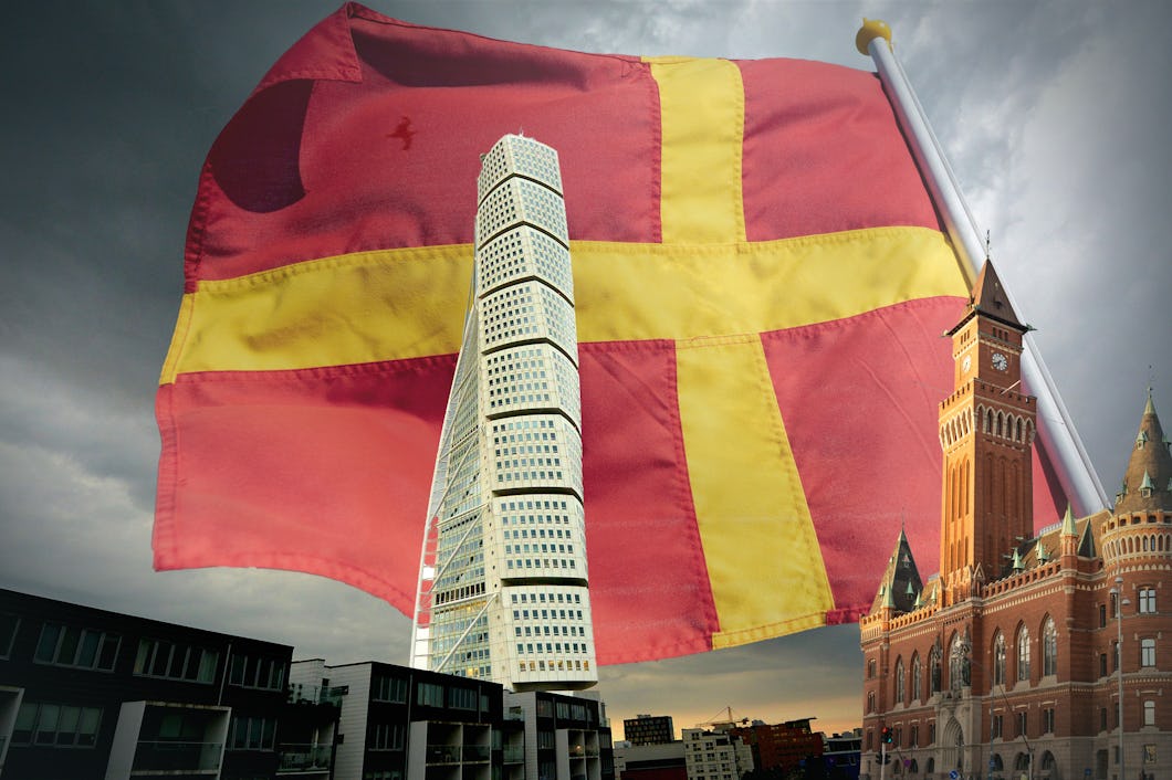 En stor röd flagga med ett gult kors vajar över ett modernt stadssilhuett med ett observatorietorn och historiska byggnader under en molnig himmel.