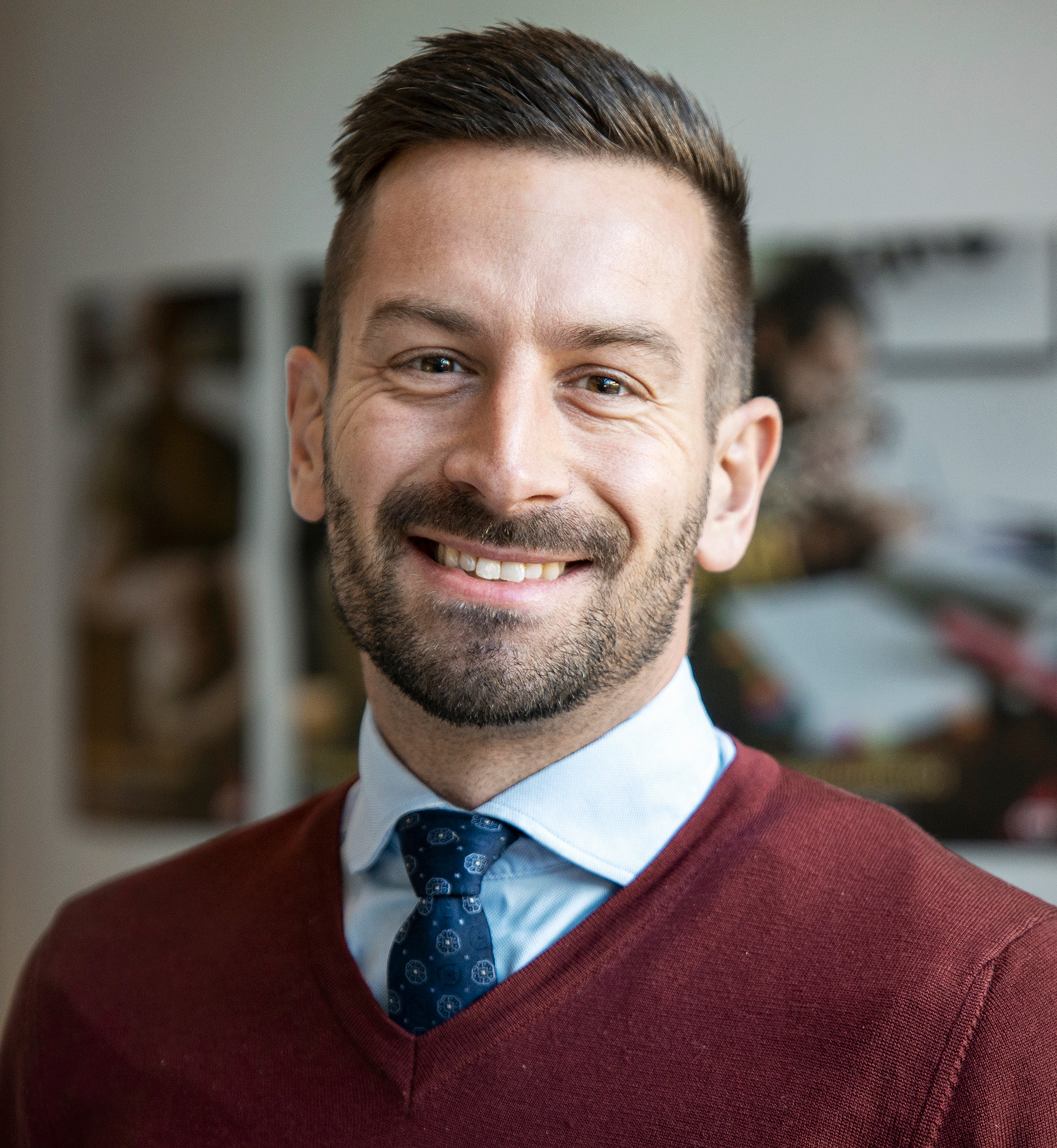 En man med kort hår och skägg står inomhus. Han bär en vinröd tröja över en ljusblå skjorta och en mönstrad blå slips. Bakgrunden är suddig med några inramade foton på väggen.