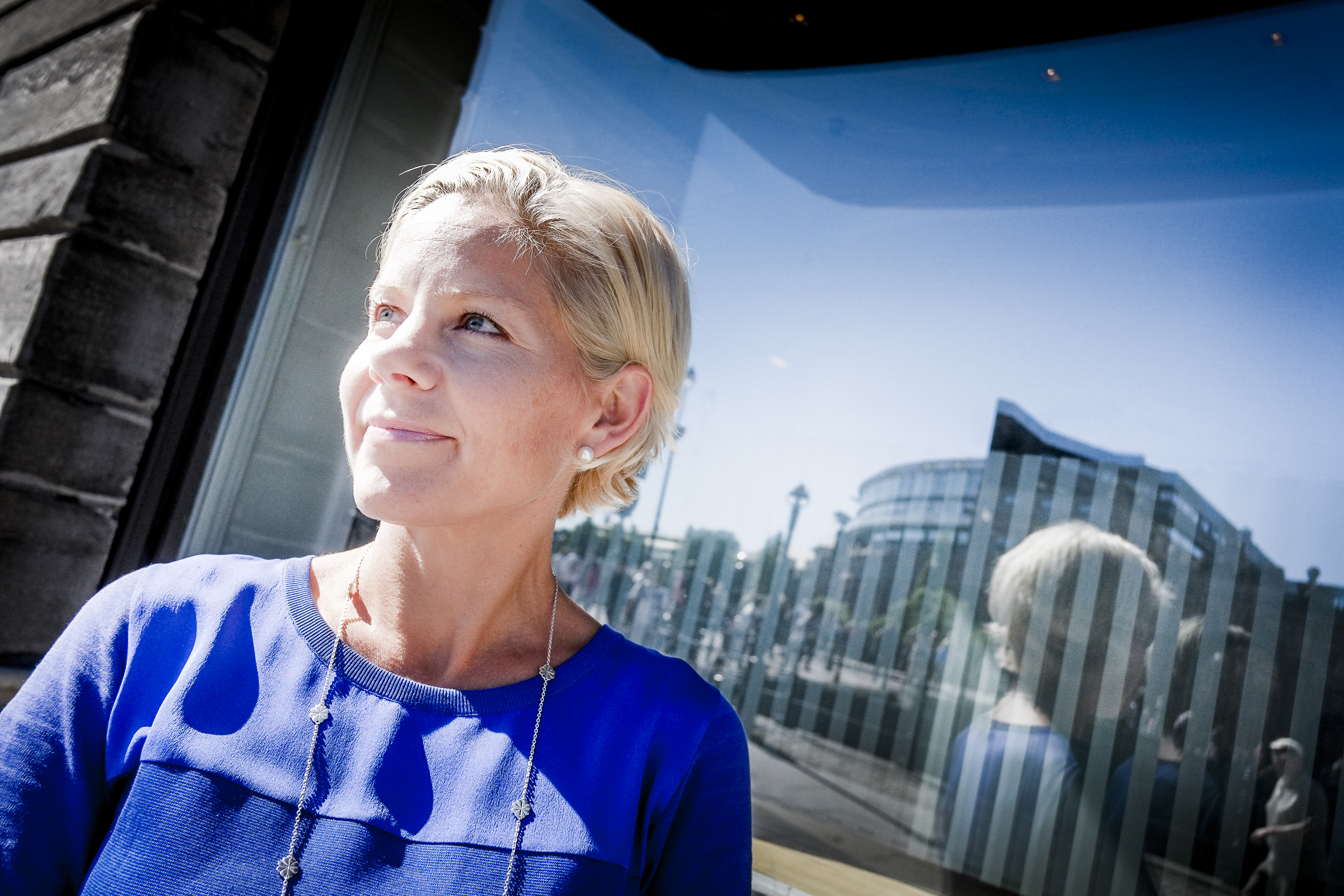 Susanna Campbell med blont hår i blå topp står framför en glasbyggnad och tittar åt sidan. I glaset bakom henne syns en spegling av stadslandskapet.