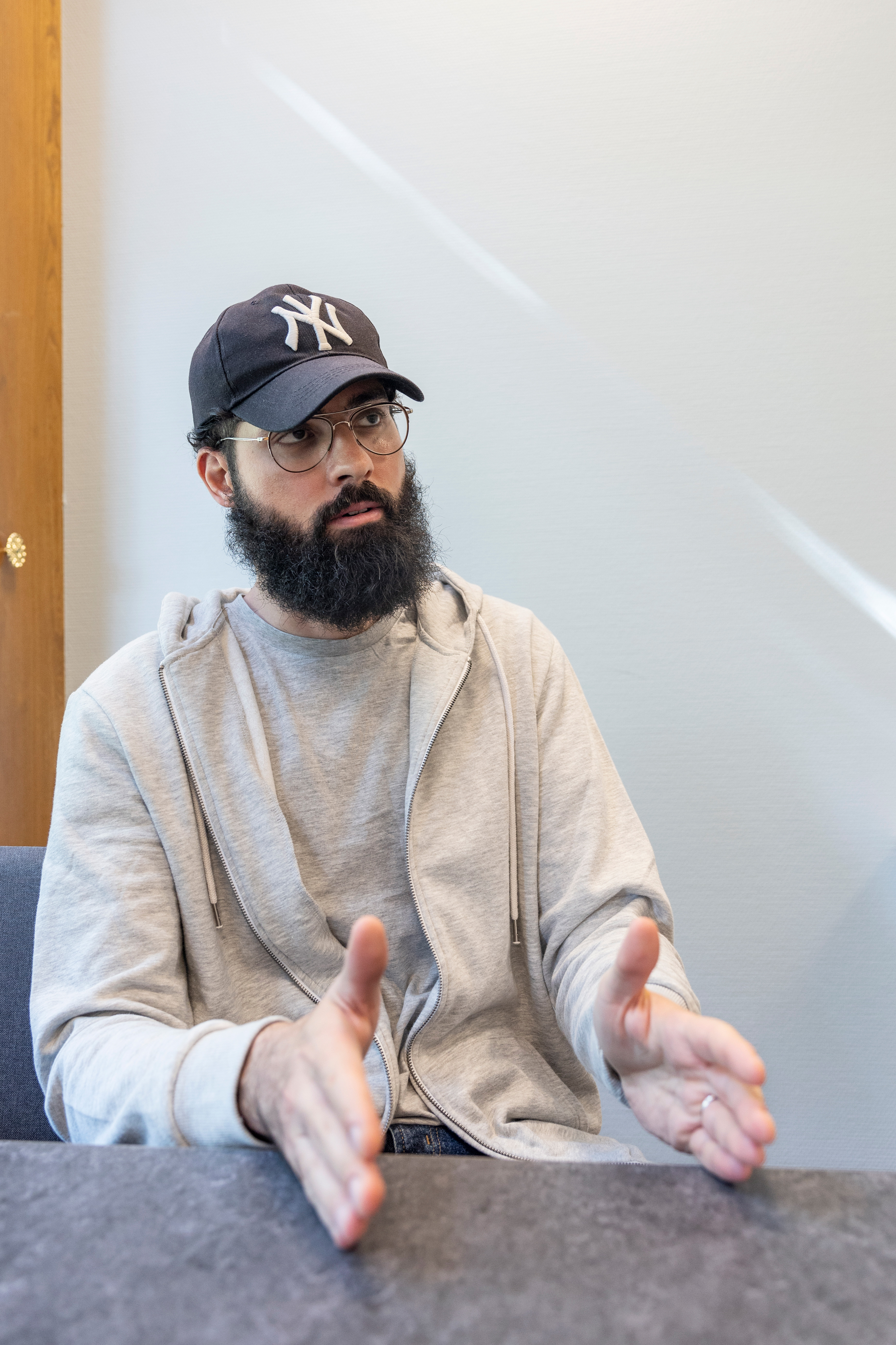 Man med skägg och glasögon som bär keps och grå huvtröja, sitter och gestikulerar med händerna medan han talar.