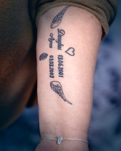 En tatuerad underarm visar text, siffror, datum, vingar, ett hjärta och en tallkotte. Tatueringen innehåller namn, datum och dekorativa symboler på insidan av armen.