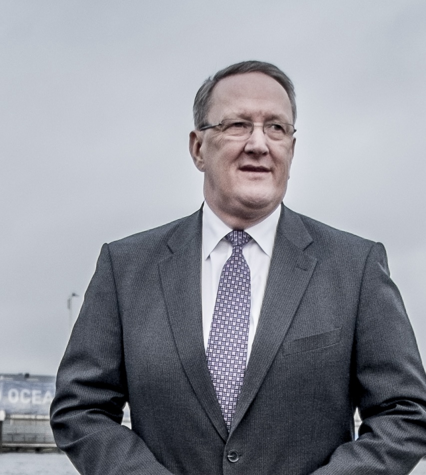 Tom Johnstone i grå kostym, vit skjorta och mönstrad slips står utomhus med en molnig himmel i bakgrunden.