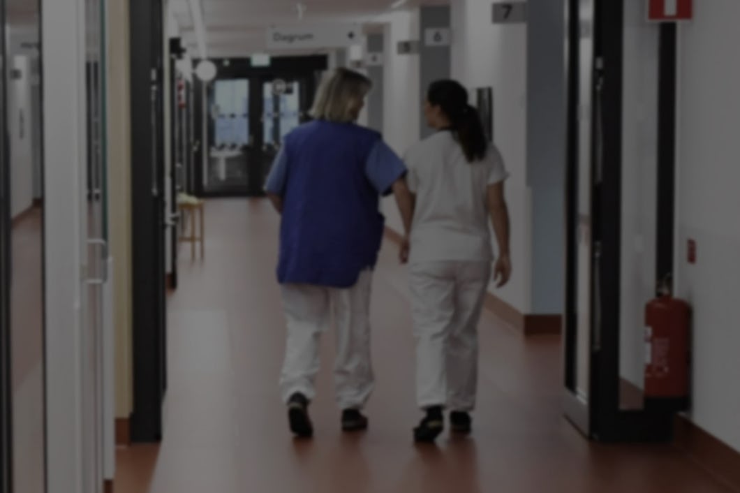 Två vårdpersonal, en i blå uniform och en i vit, går nedför en sjukhuskorridor.